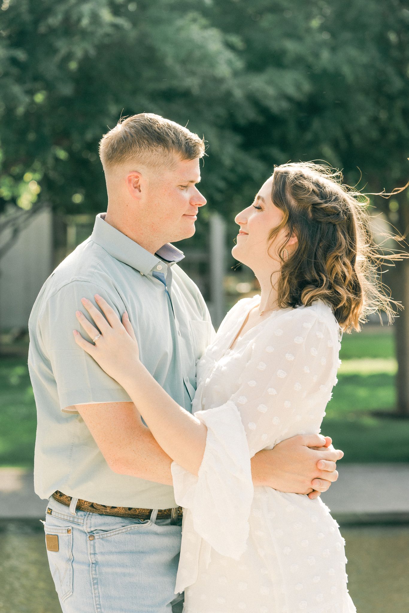 A couple in light summer attire dance and embrace joyfully in a sunlit outdoor setting with trees in the background.