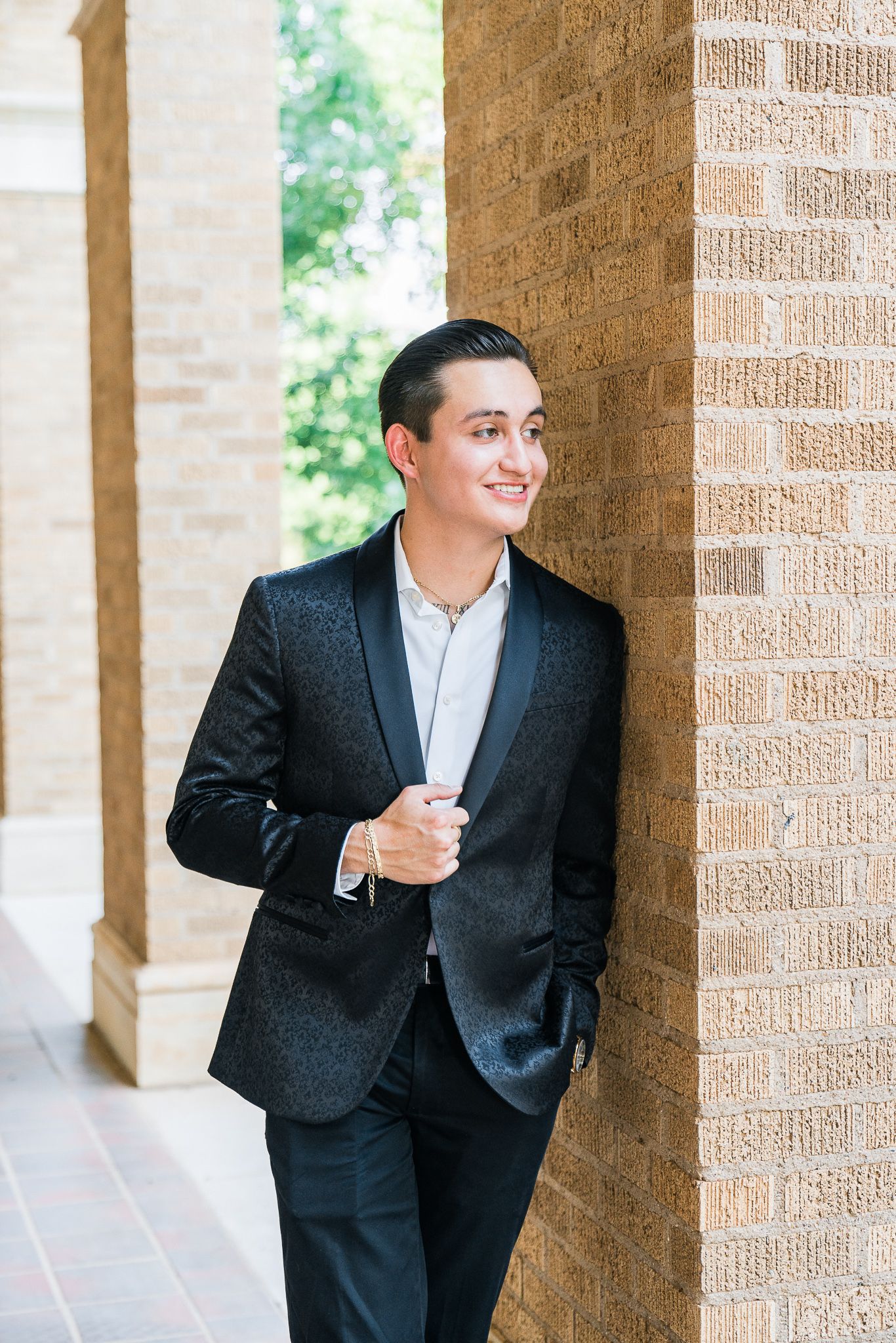 Groom in black suit laughs while leaning against brick columns in an outdoor covered walkway.