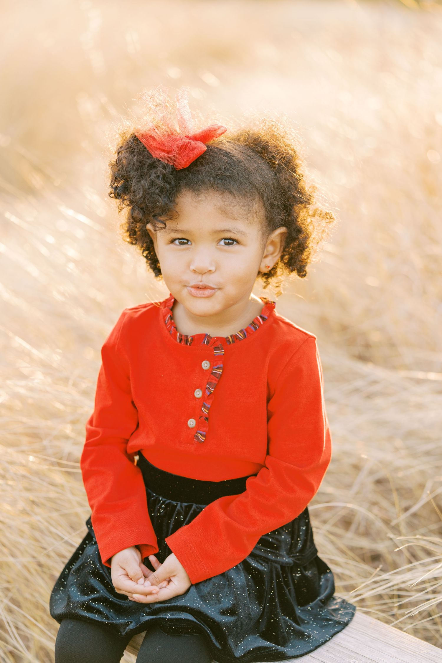 A small child in a red sweater and black skirt sits in tall grass wearing a matching red bow.
