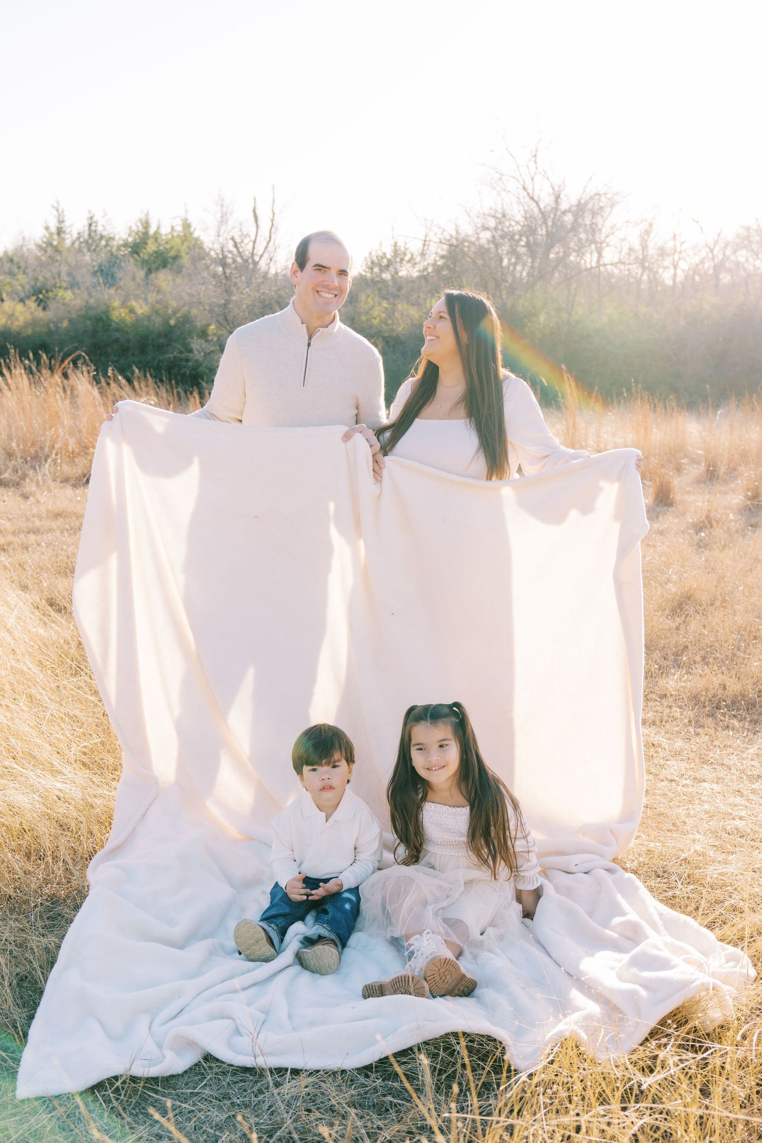 A family sits on a white blanket for a portrait session in a golden field at sunset.