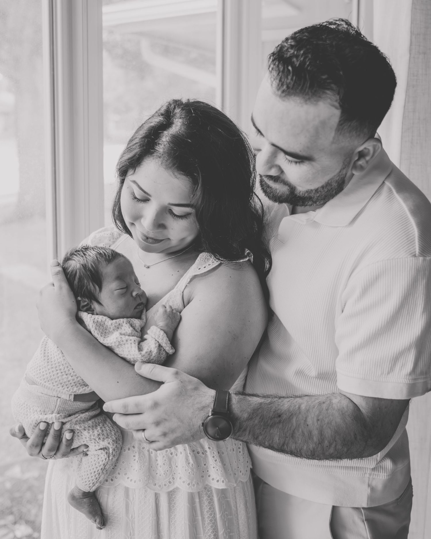 Black and white photo of new parents lovingly gazing at their newborn baby by a bright window.