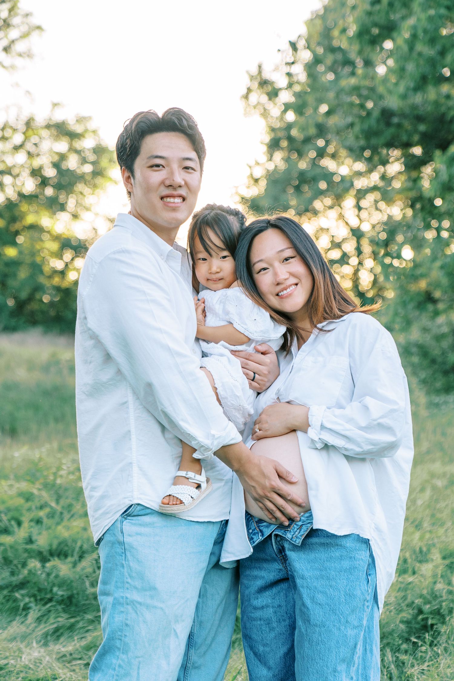 Smiling Asian family with a toddler and pregnant mother in white shirts and jeans posing outdoors in a sunlit green park.