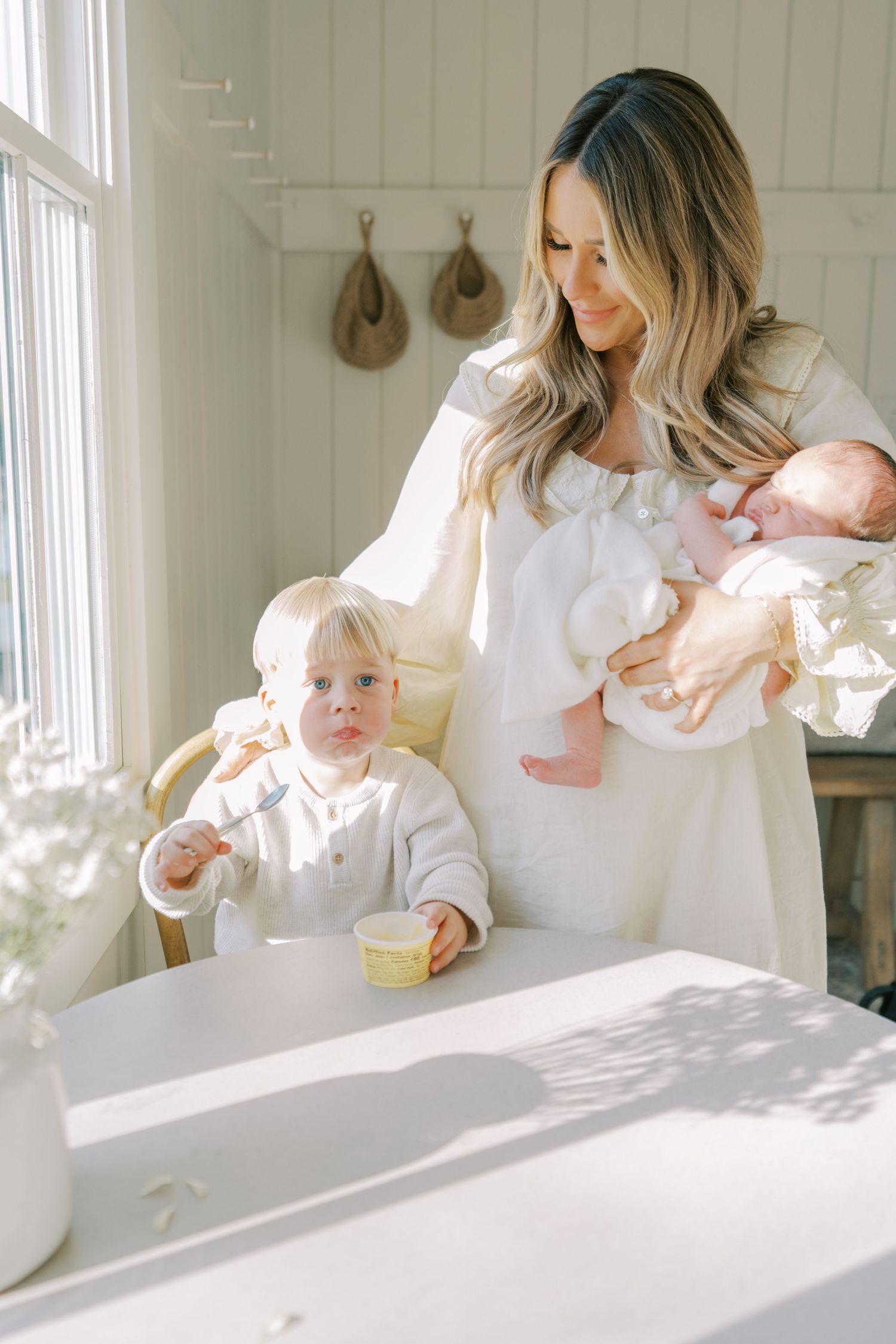 A mother in white holds a newborn while her toddler eats at a bright, airy kitchen table in a cozy home.