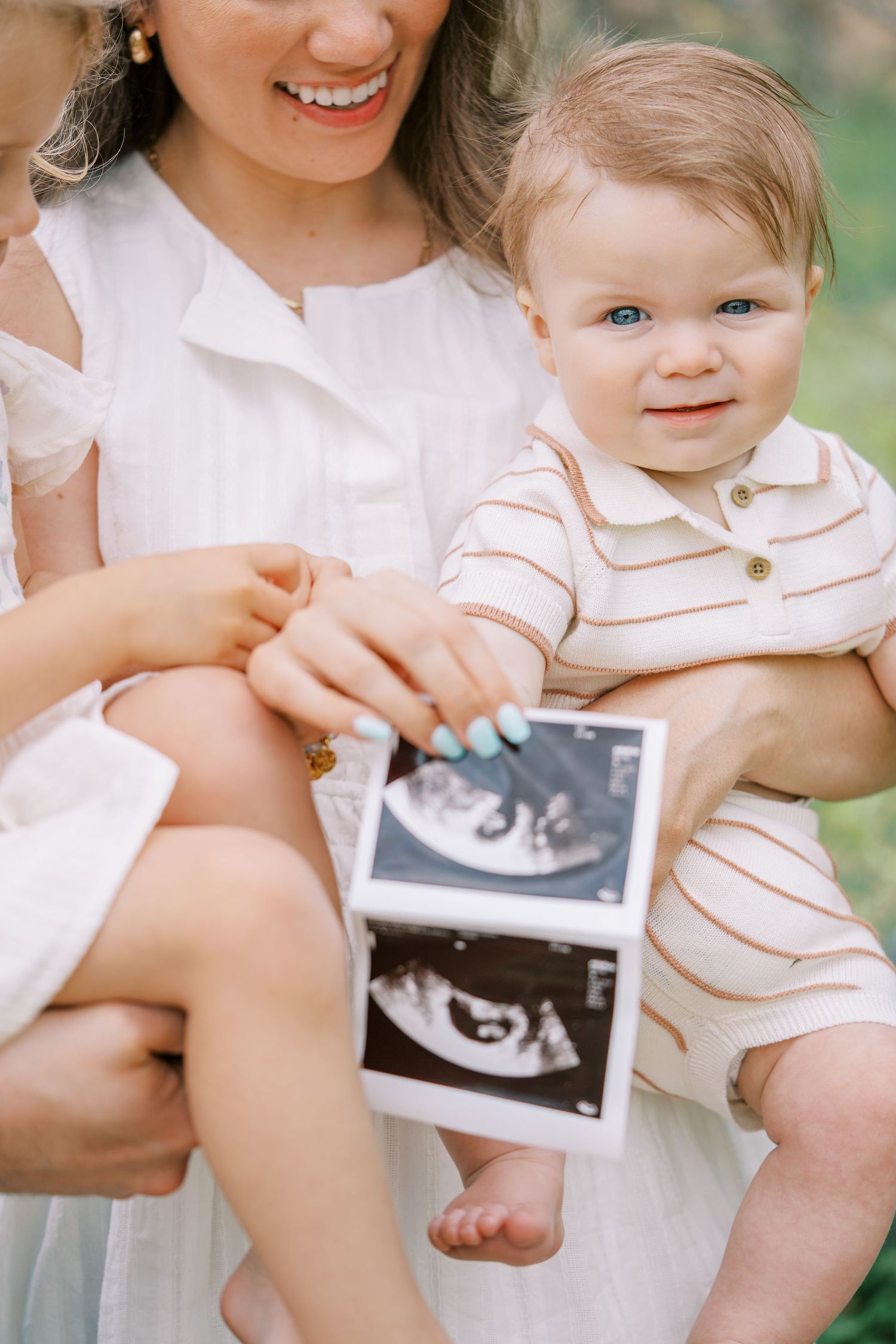 Smiling mother in white dress holds baby boy in striped romper displaying ultrasound photos outdoors.