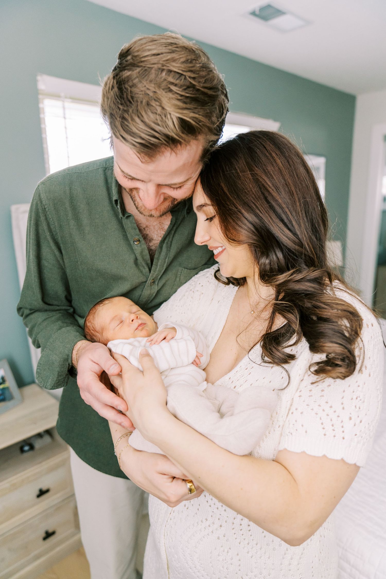 Parents share tender moments with their newborn baby in a room with mint green walls and natural lighting.
