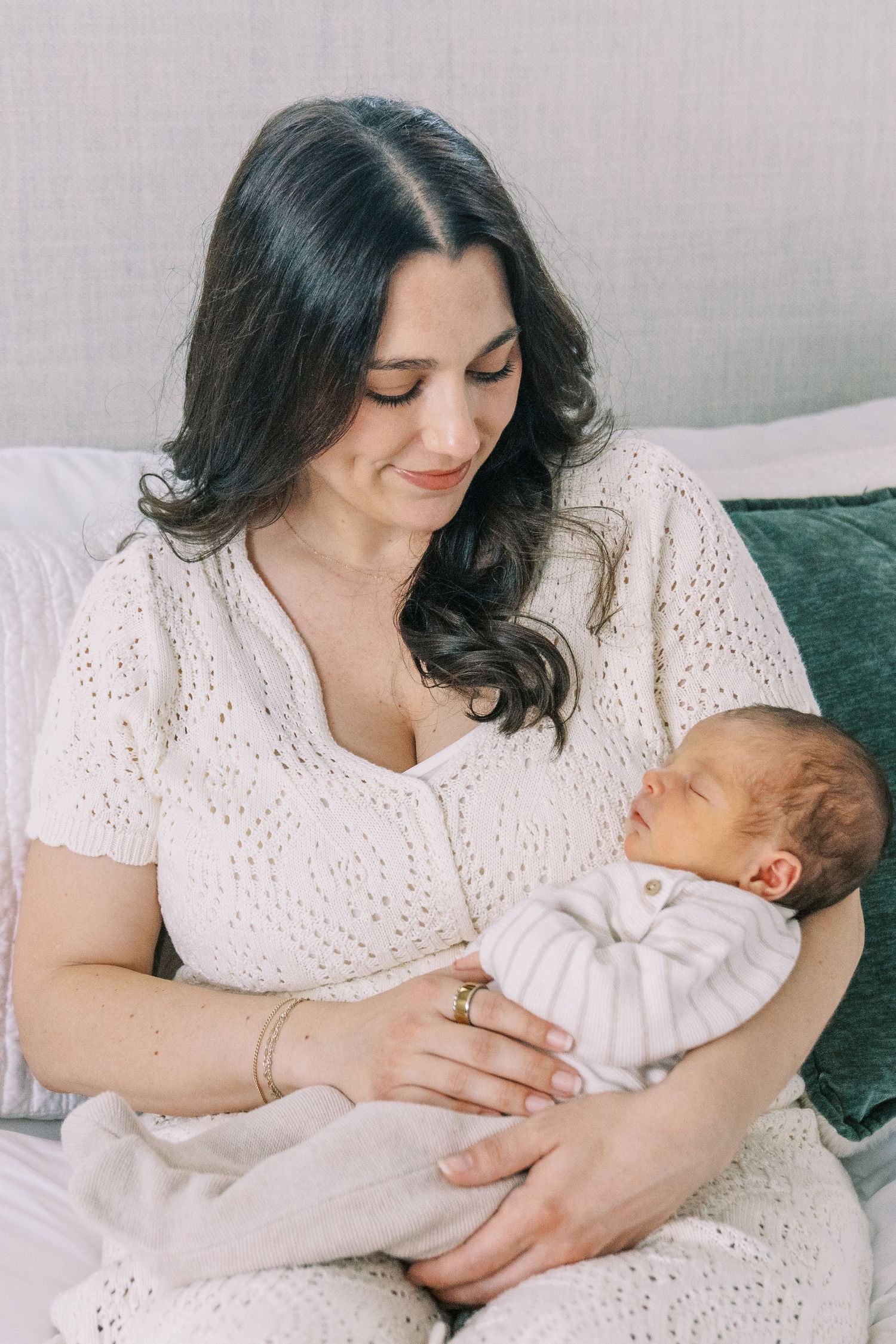 Mother in white eyelet dress cradles and nurses newborn baby in tender moment on couch.