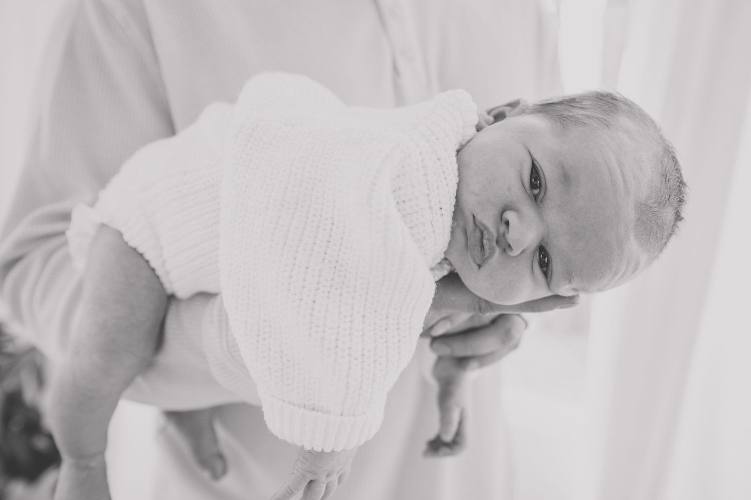 A vintage black and white photograph shows a baby in white clothing being held sideways against a light background.