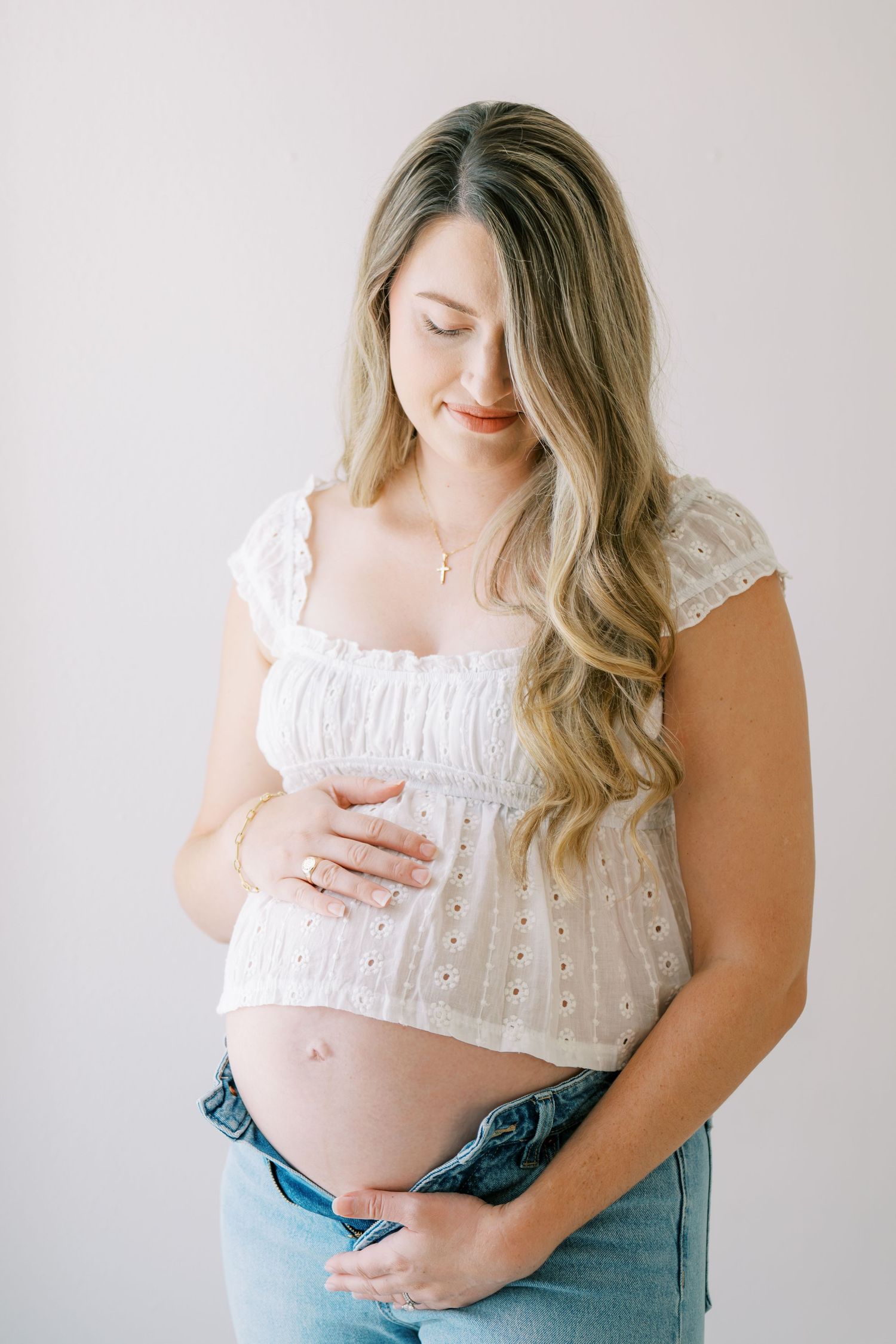 Pregnant woman in white eyelet top and jeans cradling her bare baby bump against a light background.