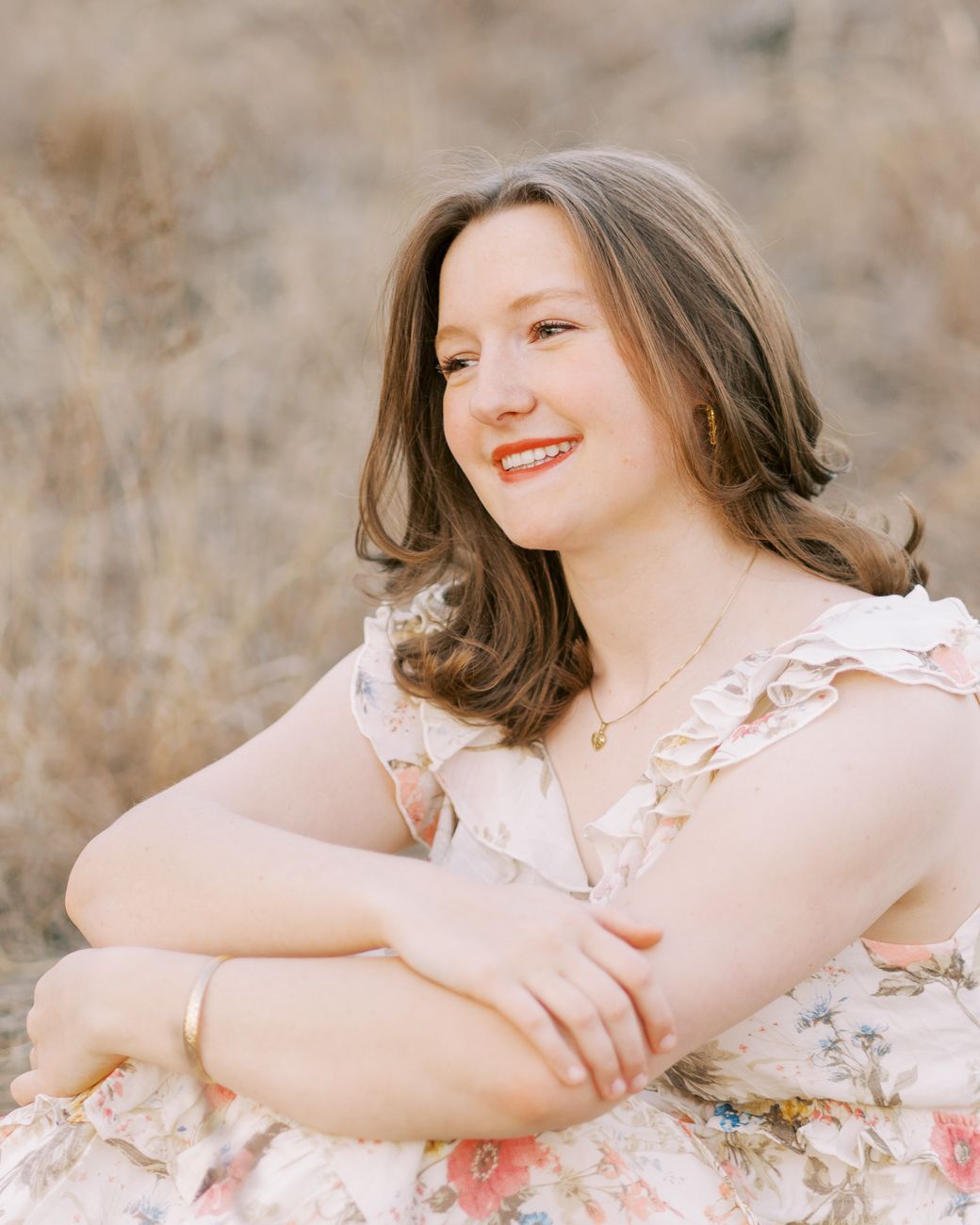 A young person in a floral dress sits in a field during golden hour with soft natural lighting.