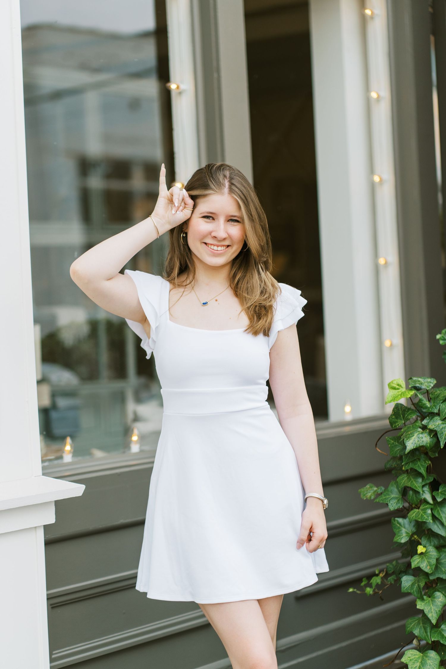 A person in a white summer dress poses cheerfully on a porch with white columns and greenery.