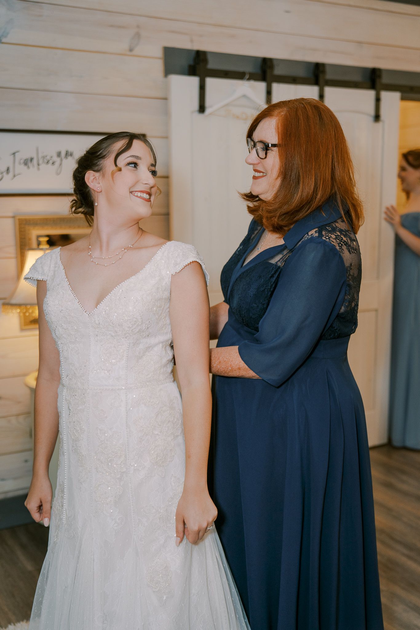 A bride in a white lace wedding dress shares a joyful moment with someone wearing a navy blue dress in a light interior space.