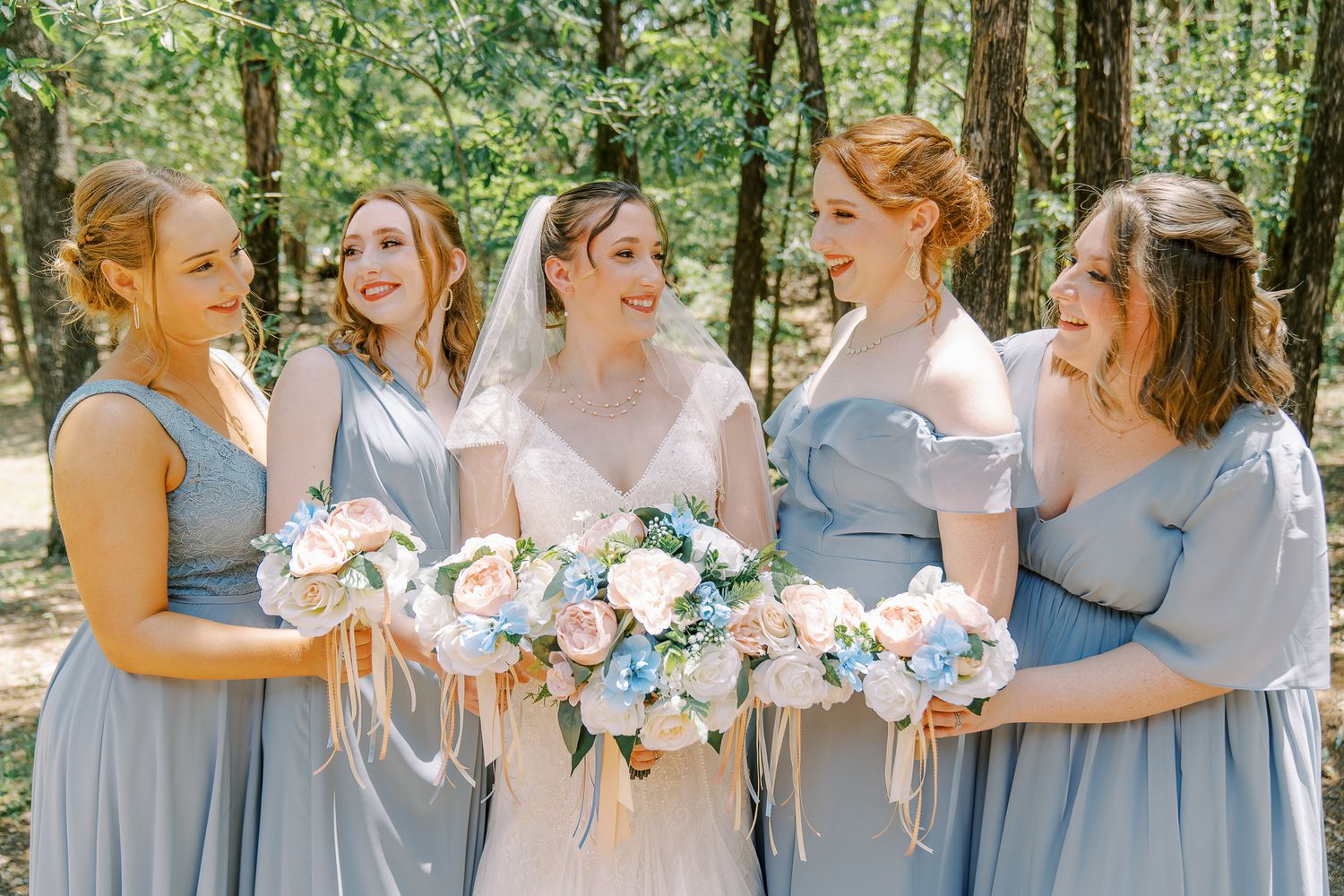 Bridesmaids in light blue dresses gather outdoors holding white floral bouquets in a woodland setting.