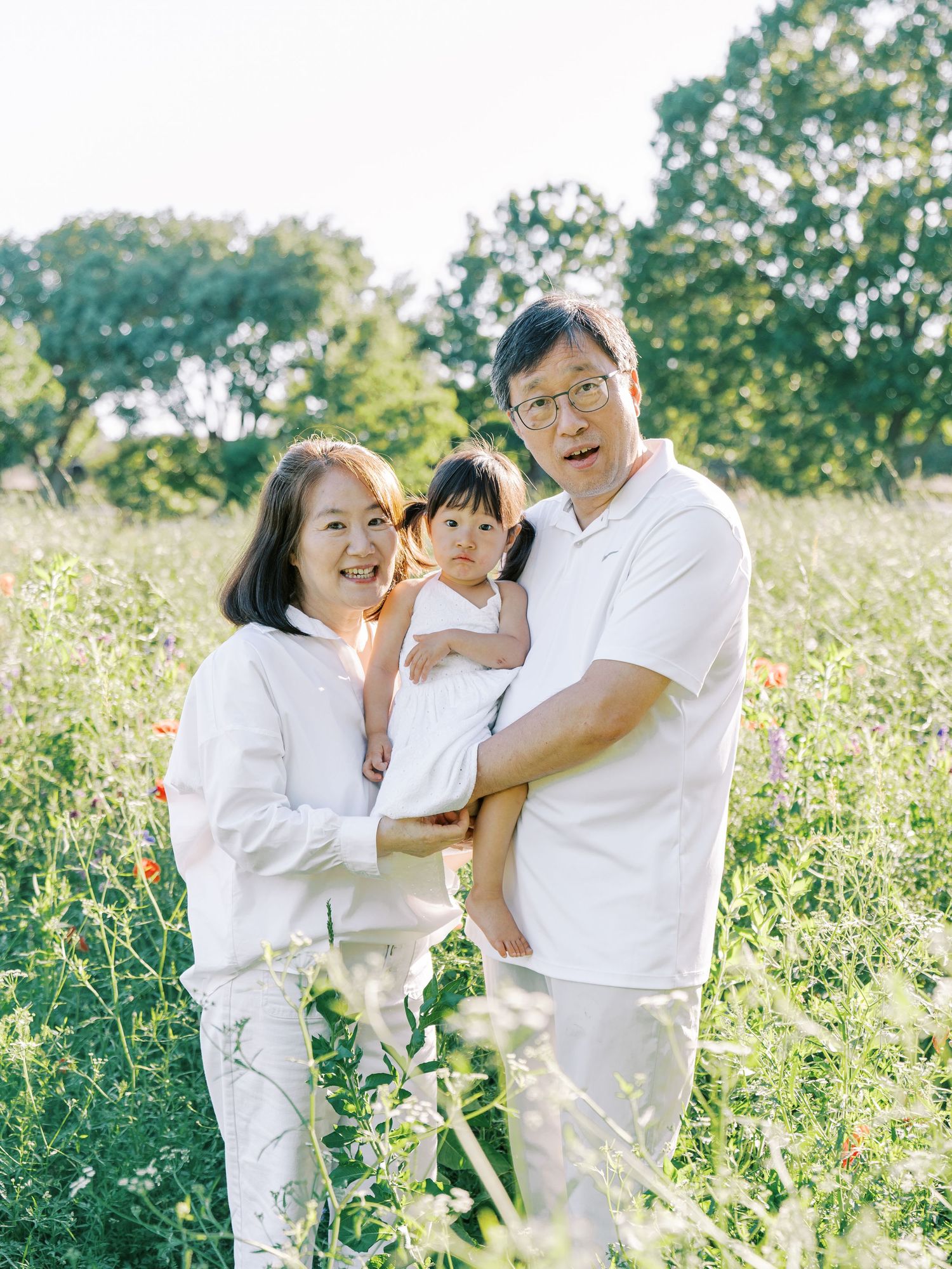 A family dressed in white outfits shares joyful moments together in a sunlit meadow filled with wildflowers.