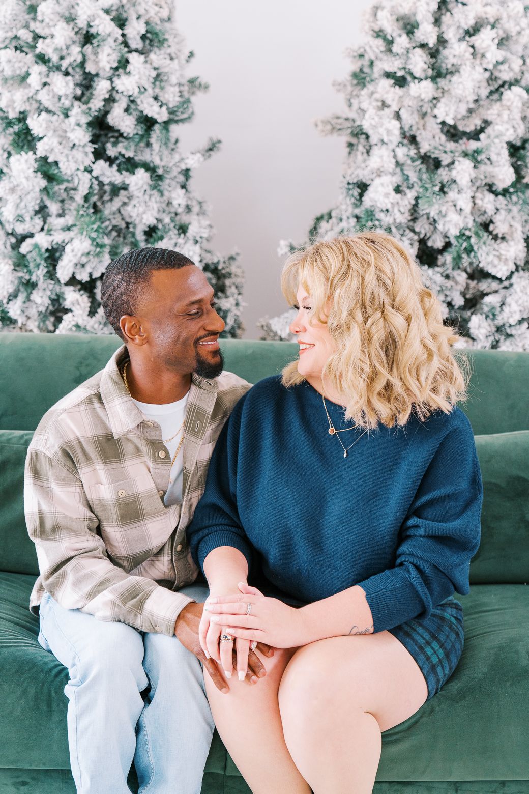 Couple sharing intimate moments on a green couch with snow-flocked Christmas trees in the background.