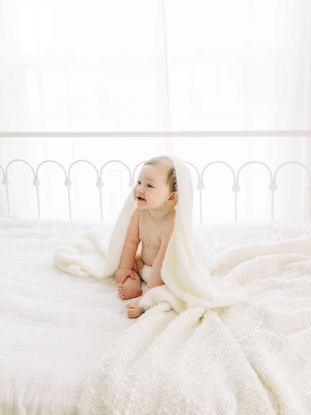 Baby playing under soft white blanket on white bedding in bright room.