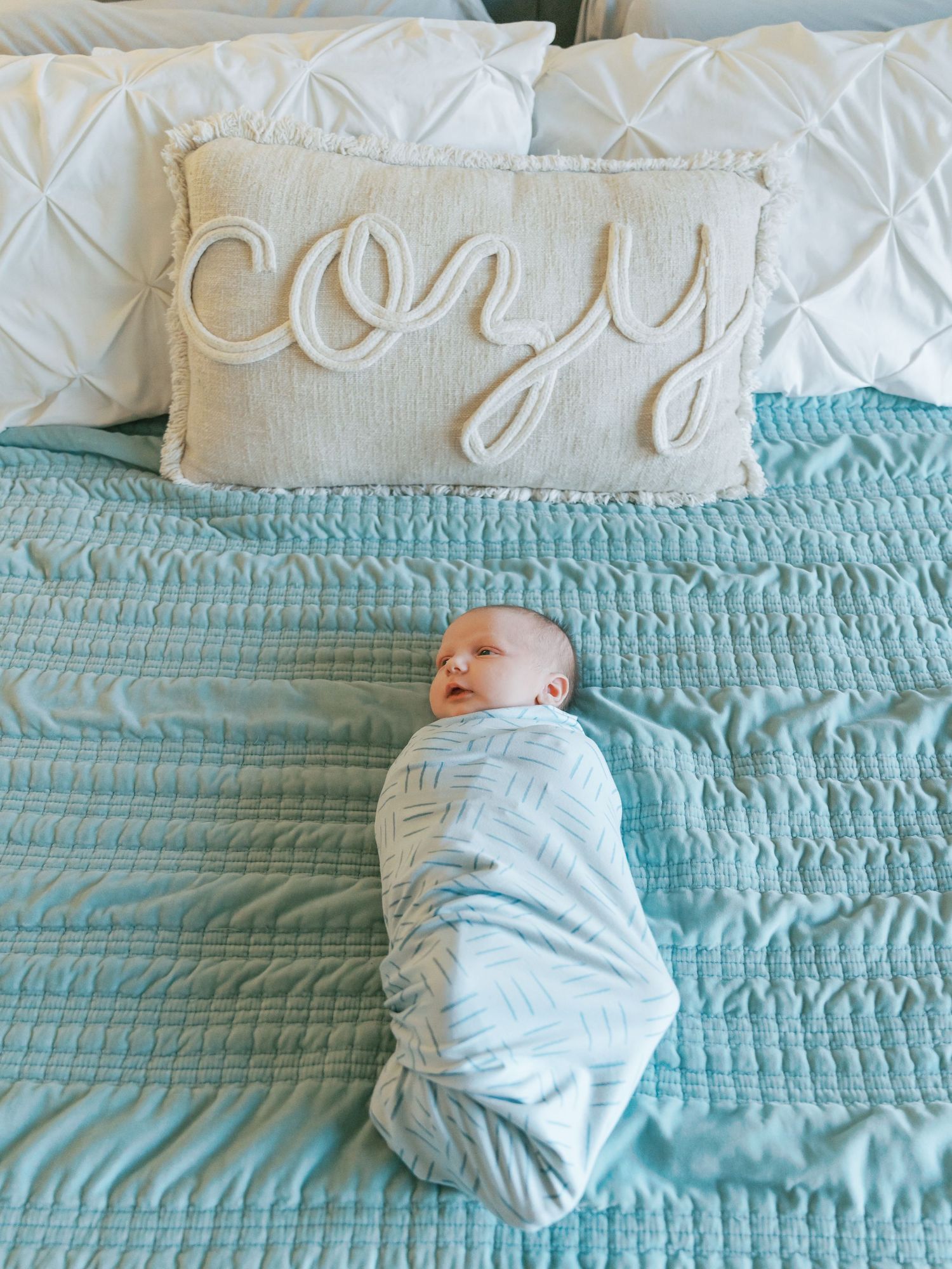Infant wrapped in light blue swaddle lies on a turquoise quilted bedspread with decorative pillows.