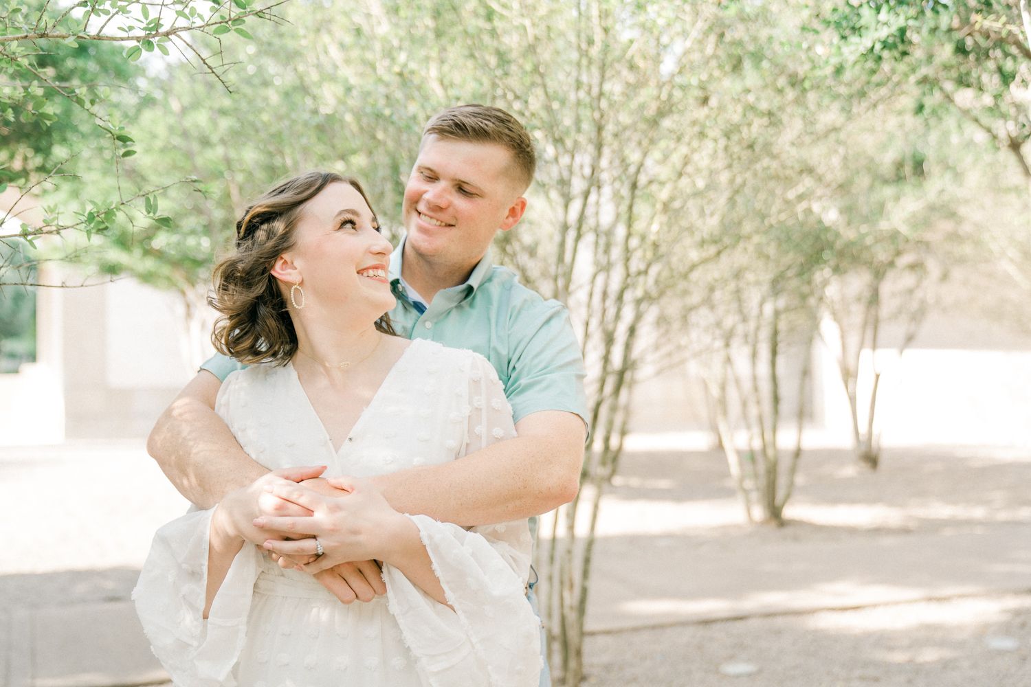 A couple shares a romantic moment in a sun-dappled outdoor setting surrounded by trees and natural light.