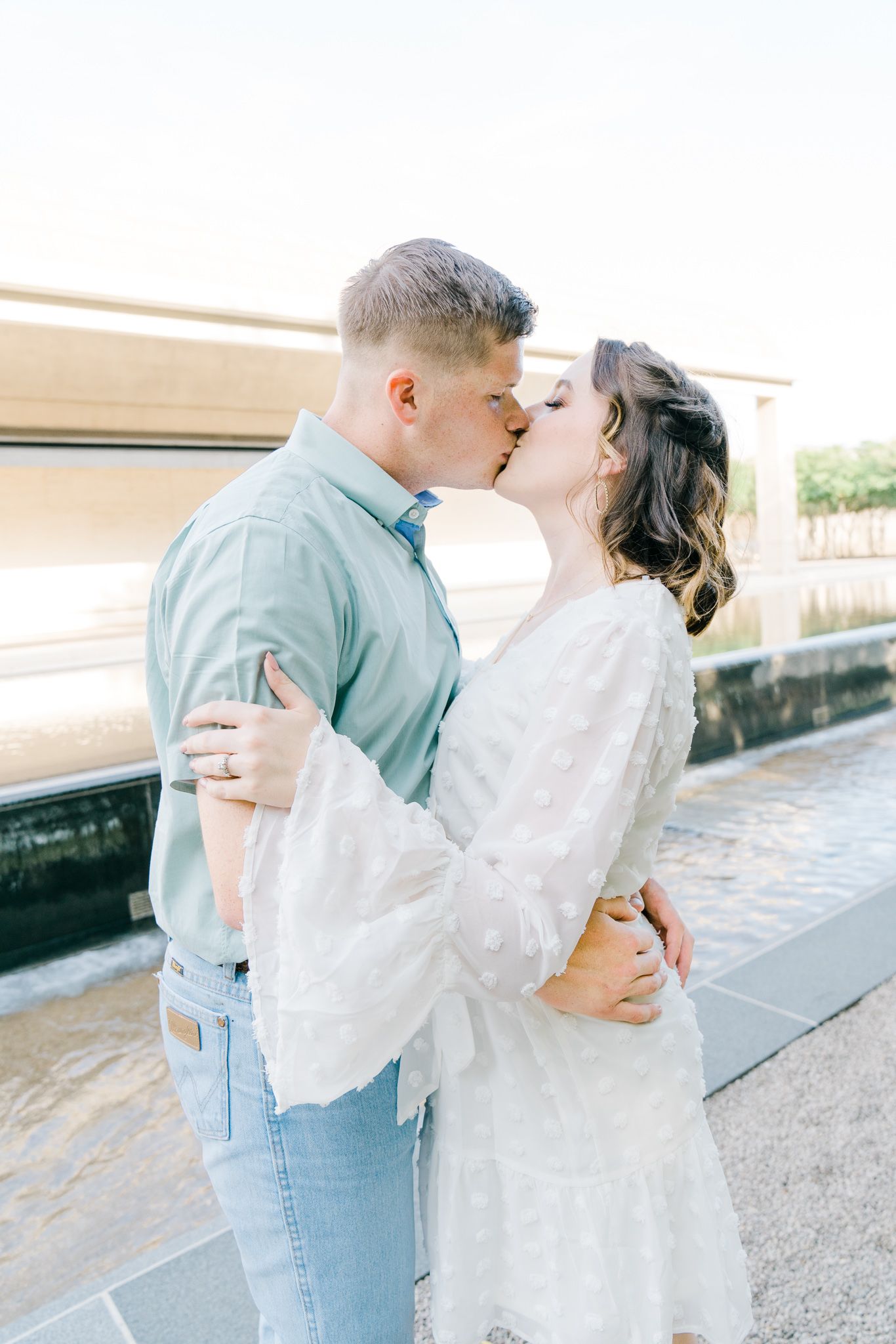 A romantic moment captured between a couple in light summer attire by a waterfront.