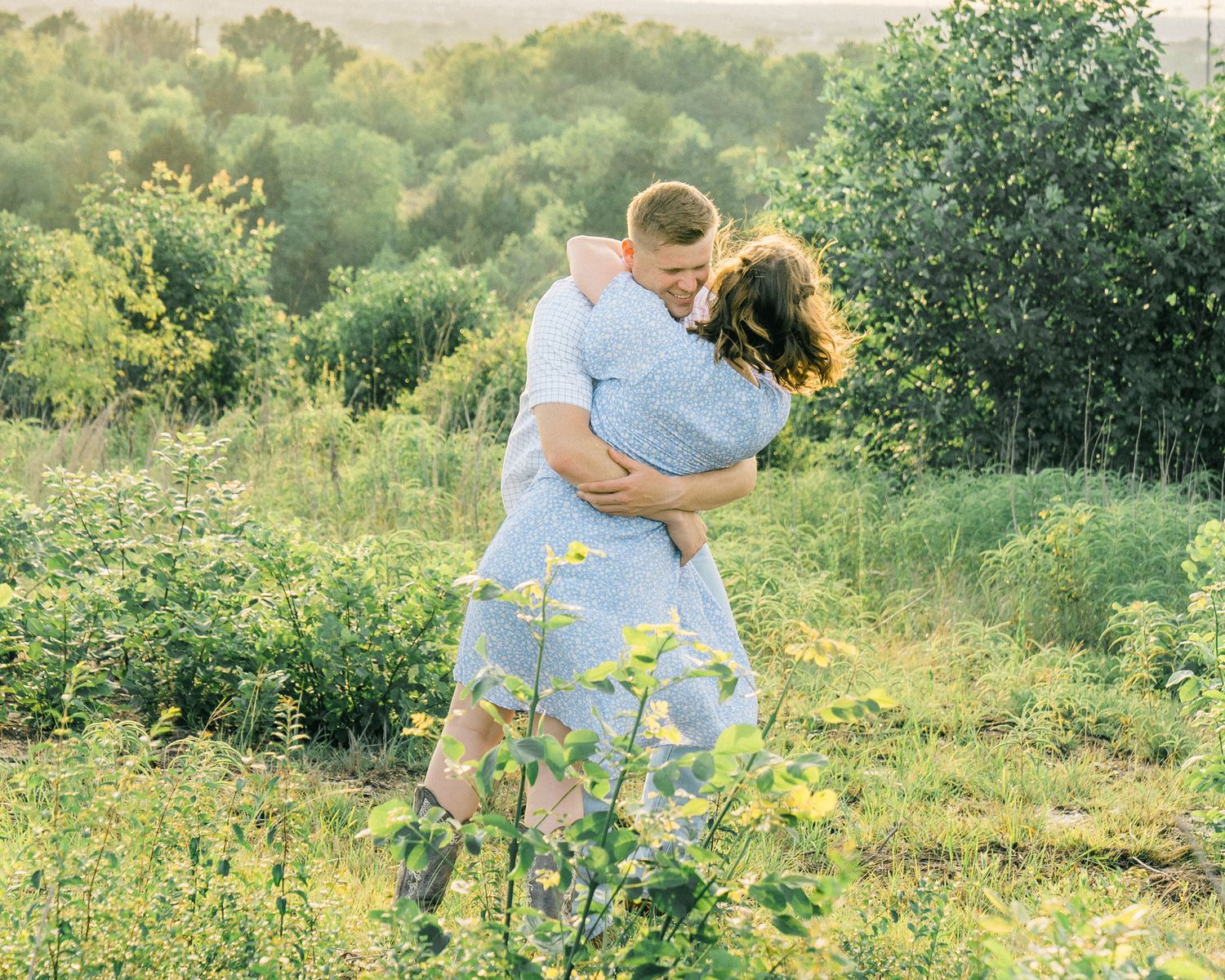 A couple embraces in a sun-drenched meadow surrounded by lush green vegetation and wildflowers.