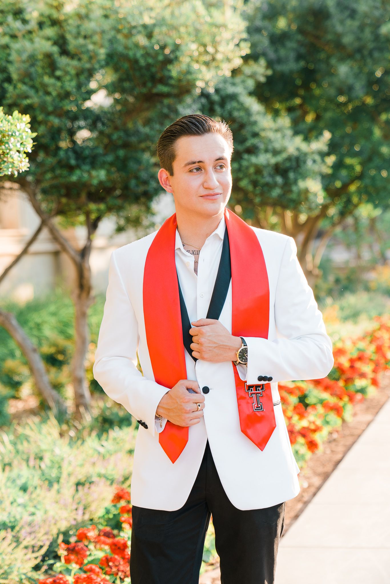 A person wearing a white blazer and red vest poses outdoors in a garden setting with trees and flowers in the background.