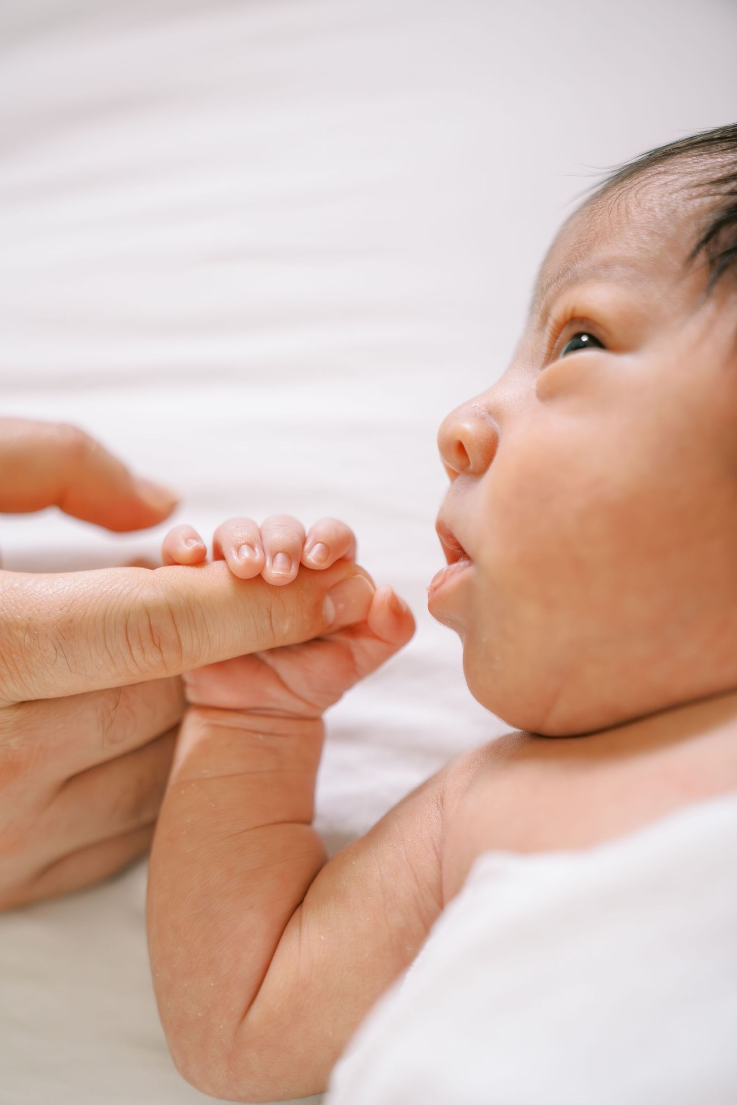 Newborn baby grasping adult finger while lying on white bed, showcasing natural bonding moment.