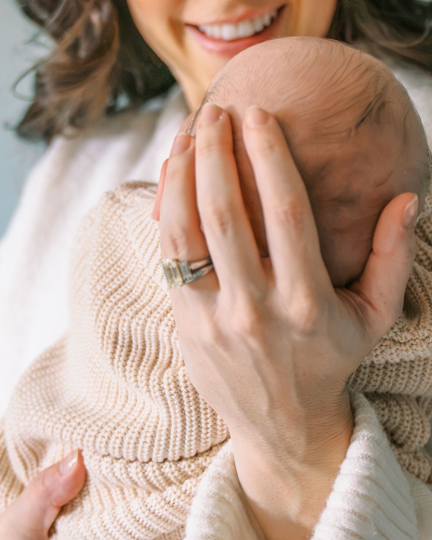 Smiling mother cradling newborn baby wrapped in soft beige knit blanket, wearing a silver ring.