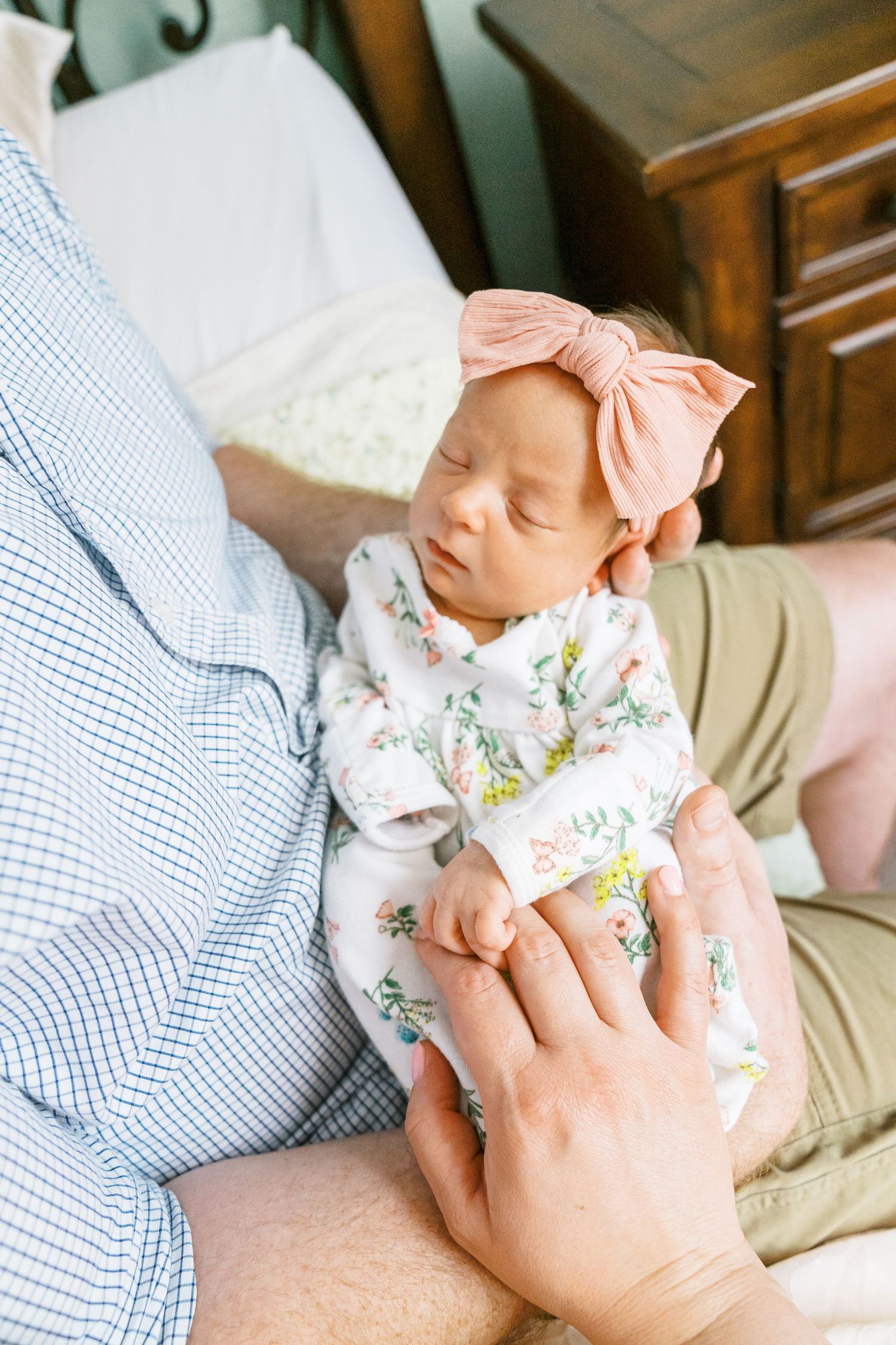 Newborn baby wearing floral outfit and pink bow rests peacefully on parent's chest.