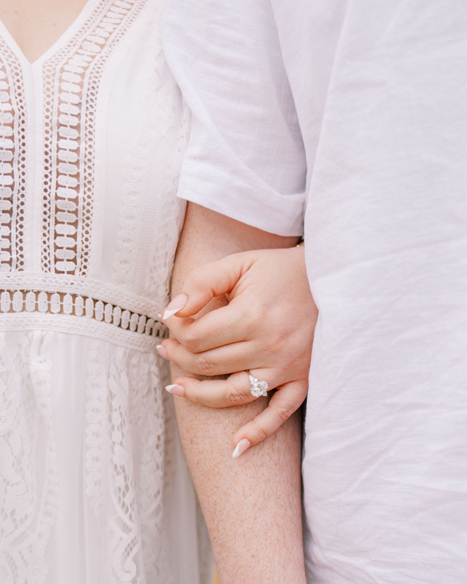 Close-up of woman wearing large diamond engagement ring holding her partners arm in white lace dress.