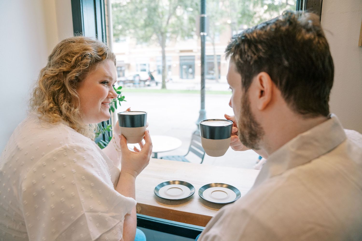 Couple sharing coffee together at a bright cafe window table during an engagement photo session.