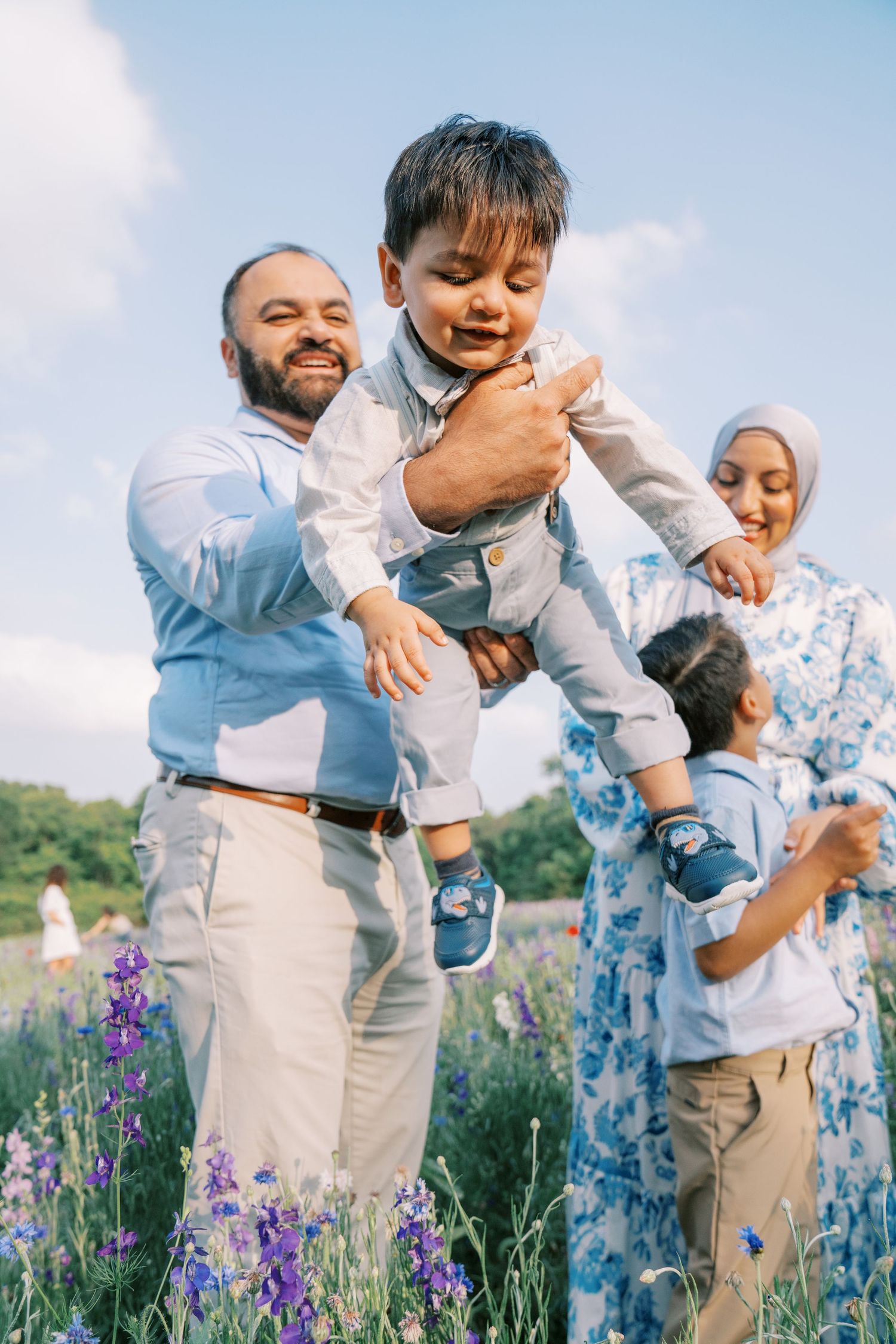 Happy Muslim family playing outdoors in a flower field, father lifting young son while mother and child smile nearby.
