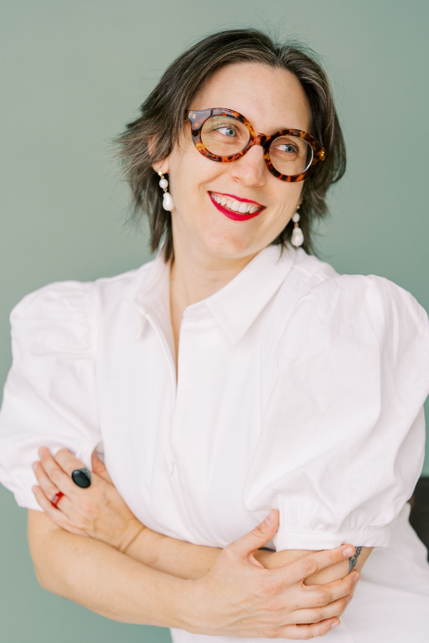Stylish portrait in a white puff-sleeve blouse with tortoise glasses and statement earrings against a mint wall.