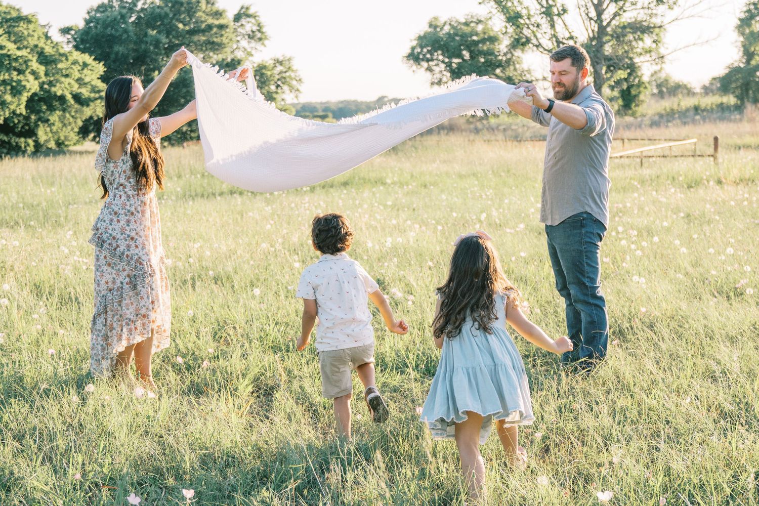 Family of four playing with white blanket in sunny green field, parents and two children enjoying outdoor fun.