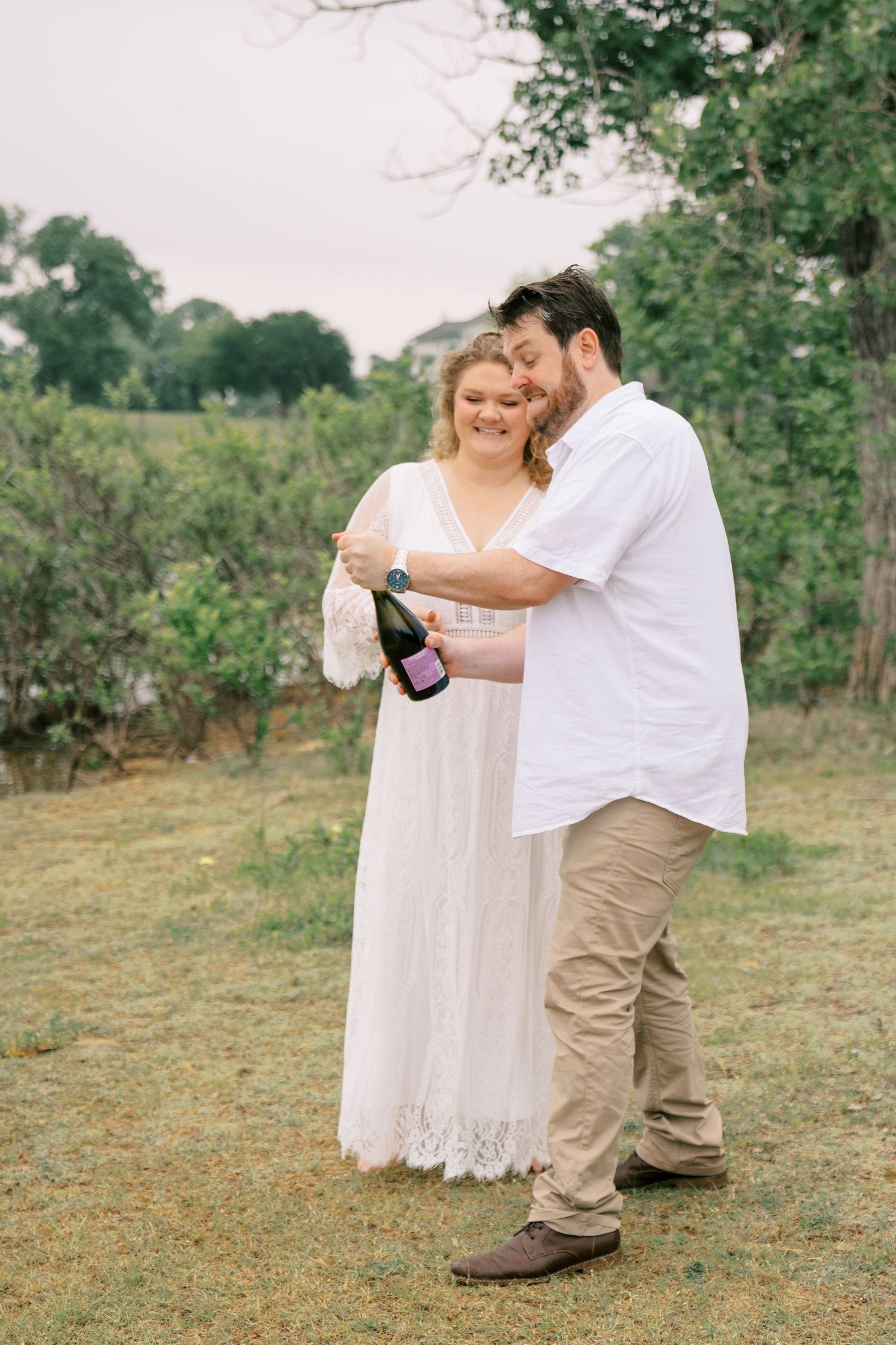 Couple in white celebrating outdoors as groom opens a bottle of champagne near trees.