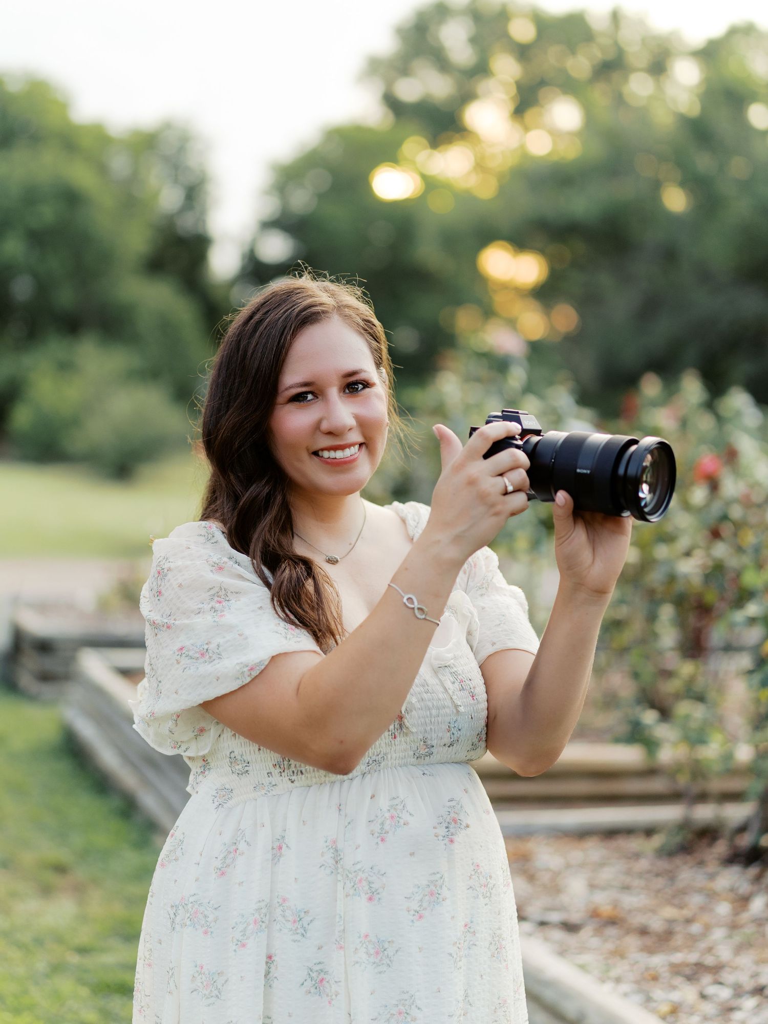 Smiling woman in white floral dress holding a DSLR camera outdoors in a lush green garden setting.