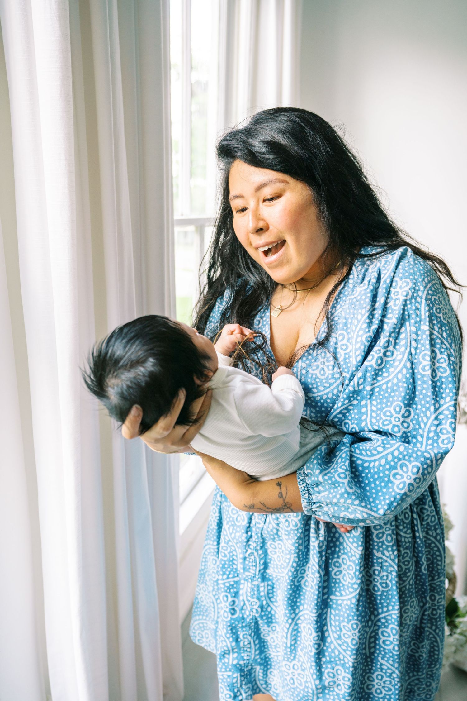 A smiling woman in a blue dress lovingly holds her newborn baby near a bright window.