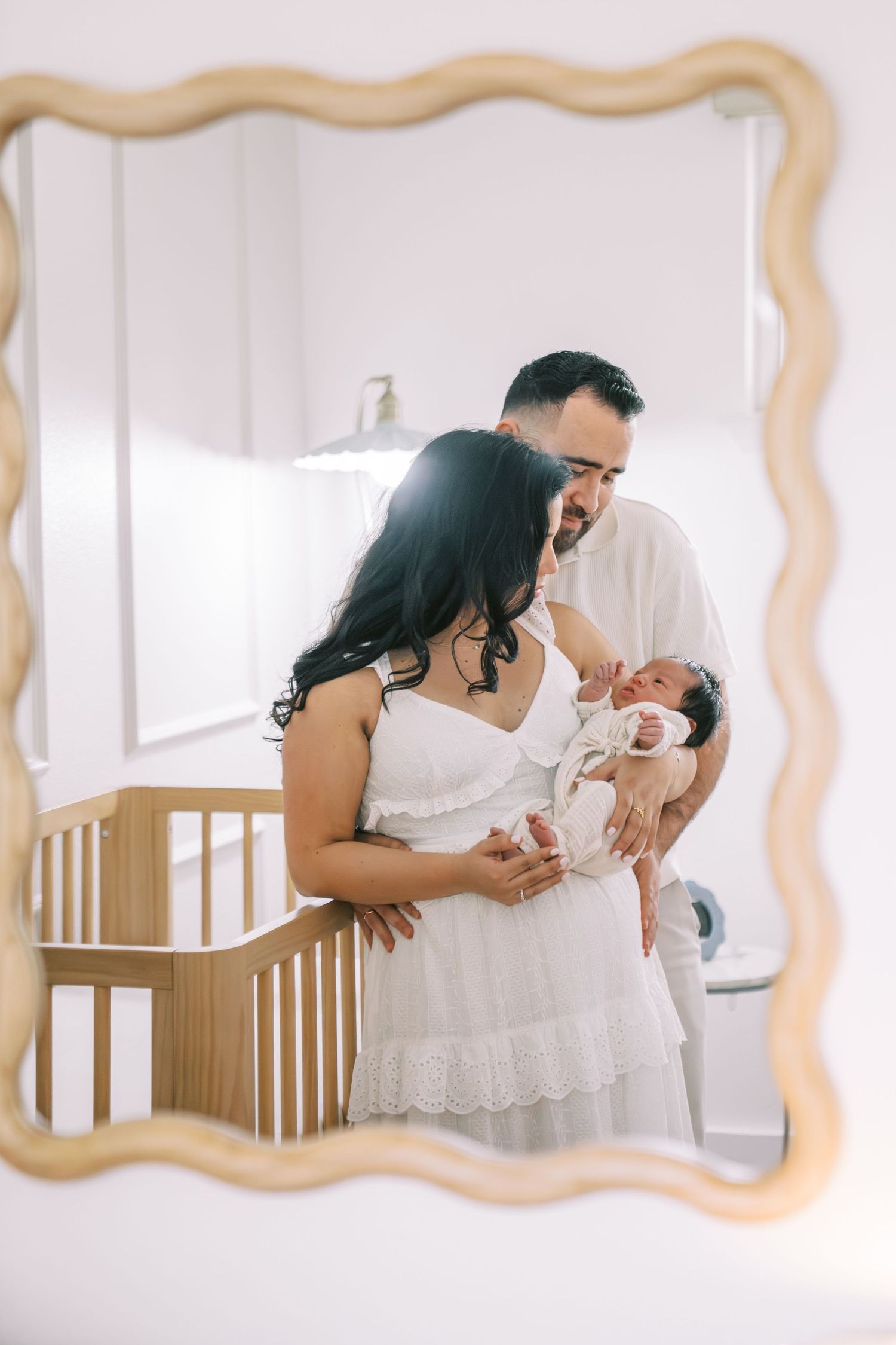 New parents holding newborn baby in nursery, reflected in ornate gold mirror, wearing white outfits.