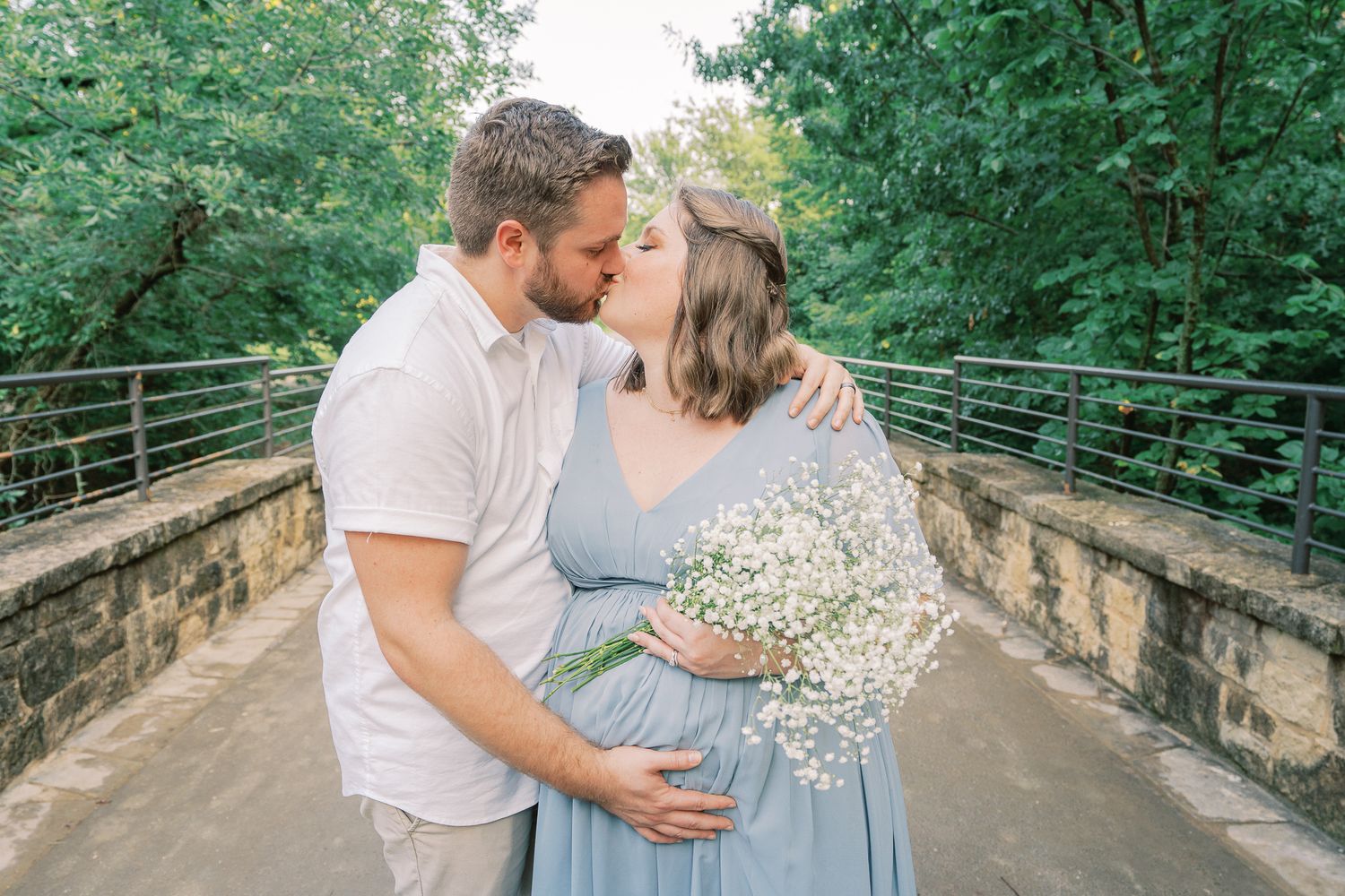 An intimate moment between an expecting couple sharing a kiss on a stone bridge surrounded by greenery.