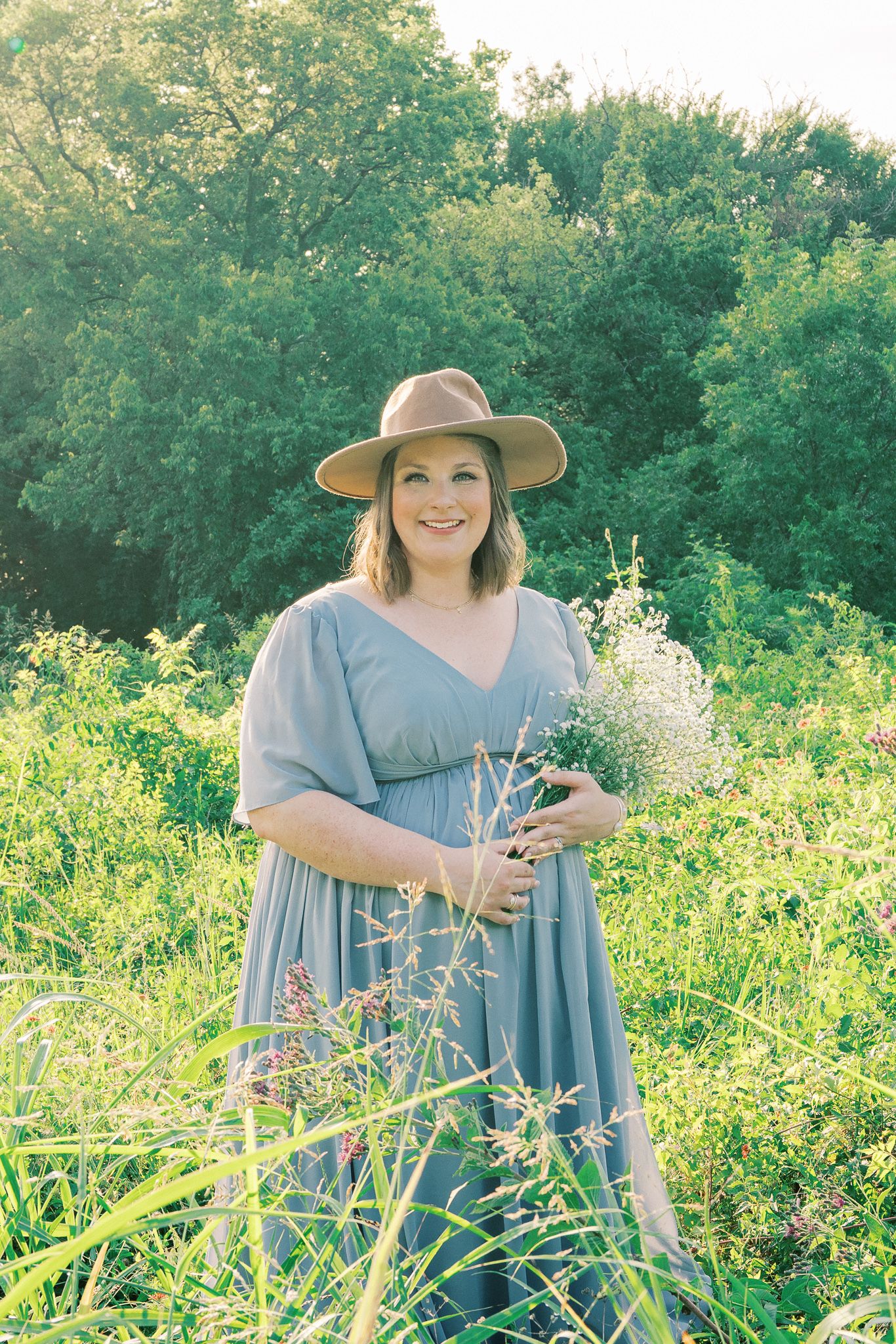 A person in a blue dress stands among wildflowers in a sunlit field wearing a sun hat.
