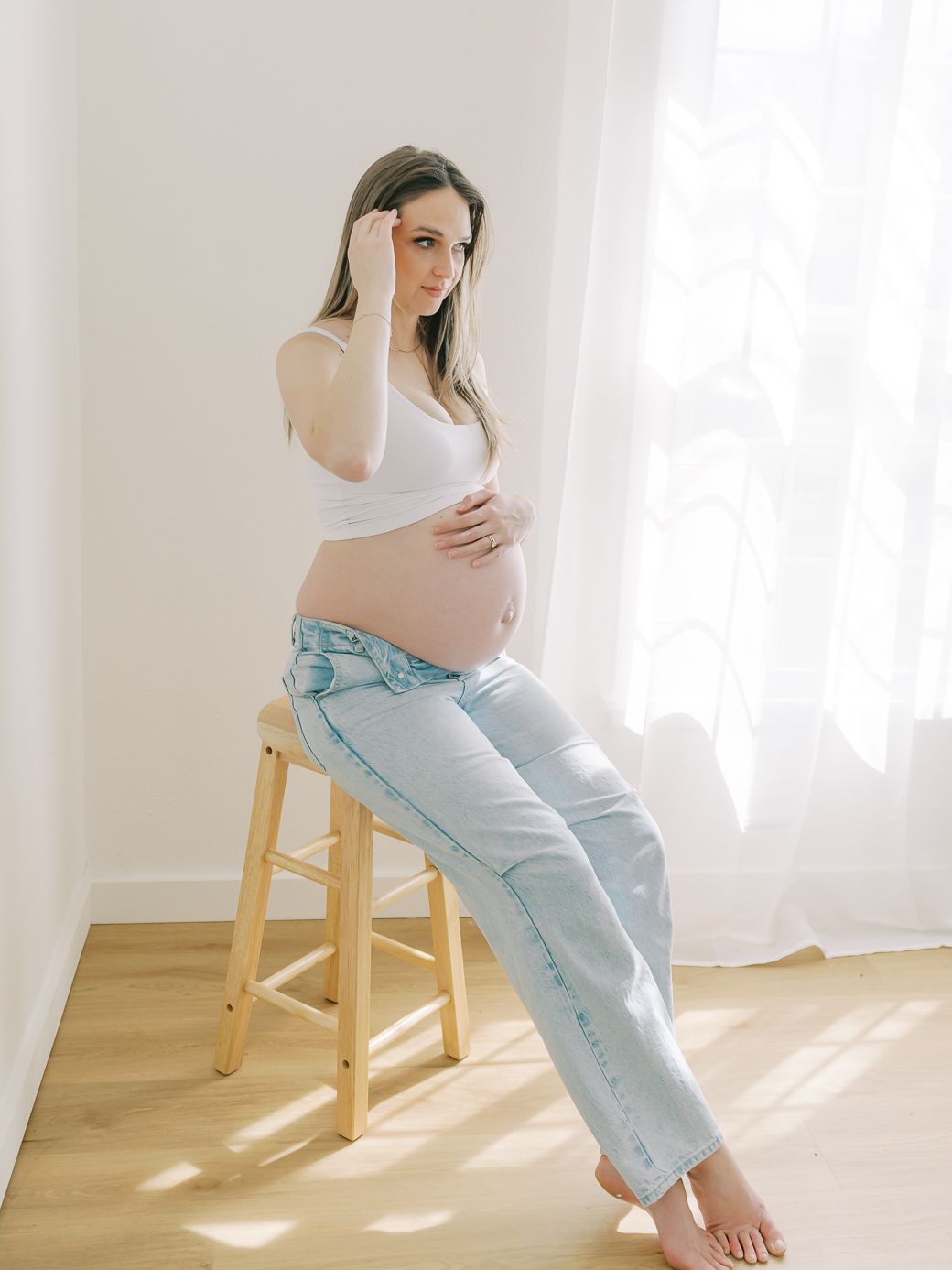 Expecting mother in white top and jeans sitting on wooden stool by window.