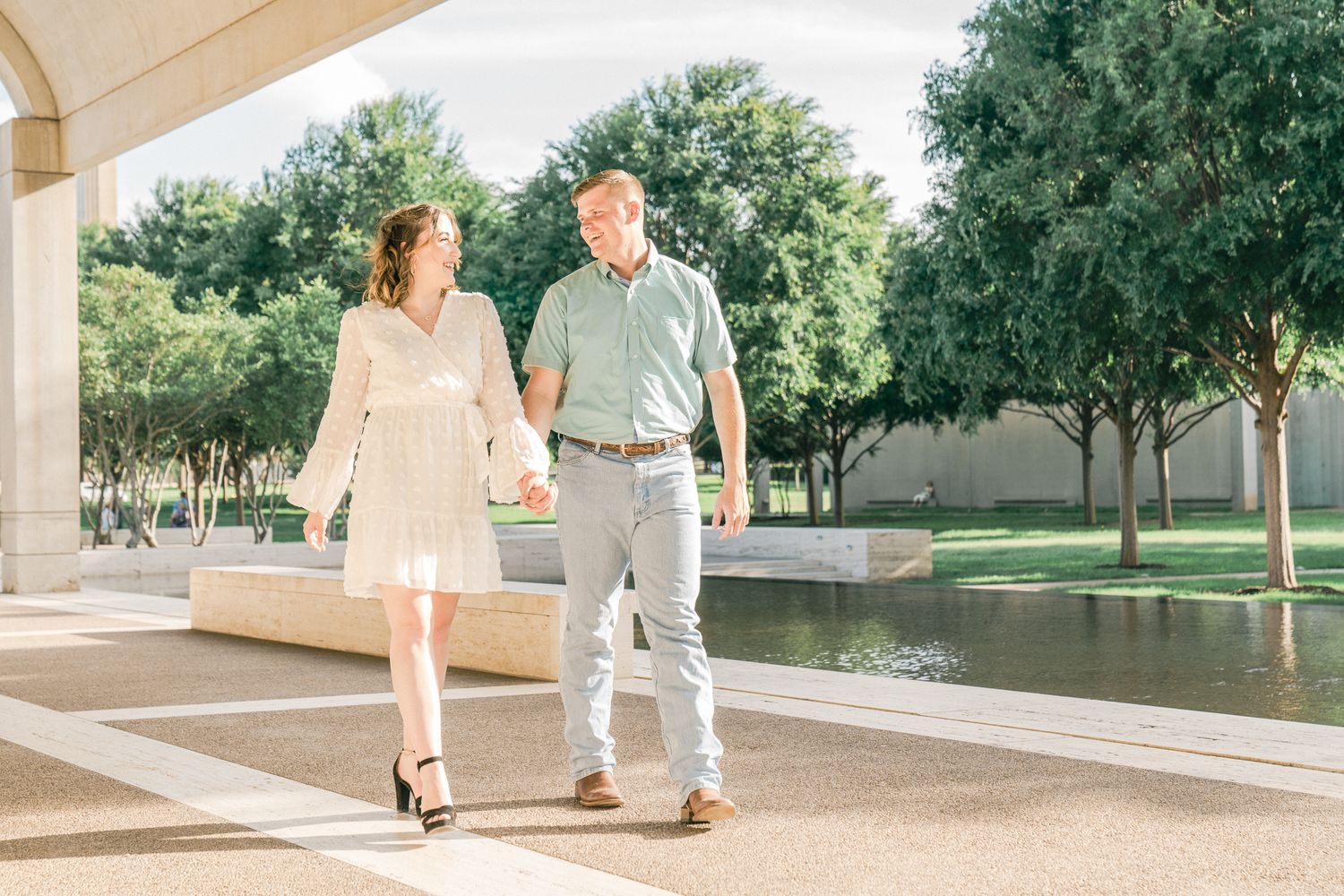 Romantic couple strolling under an architectural colonnade with trees and water in the background.
