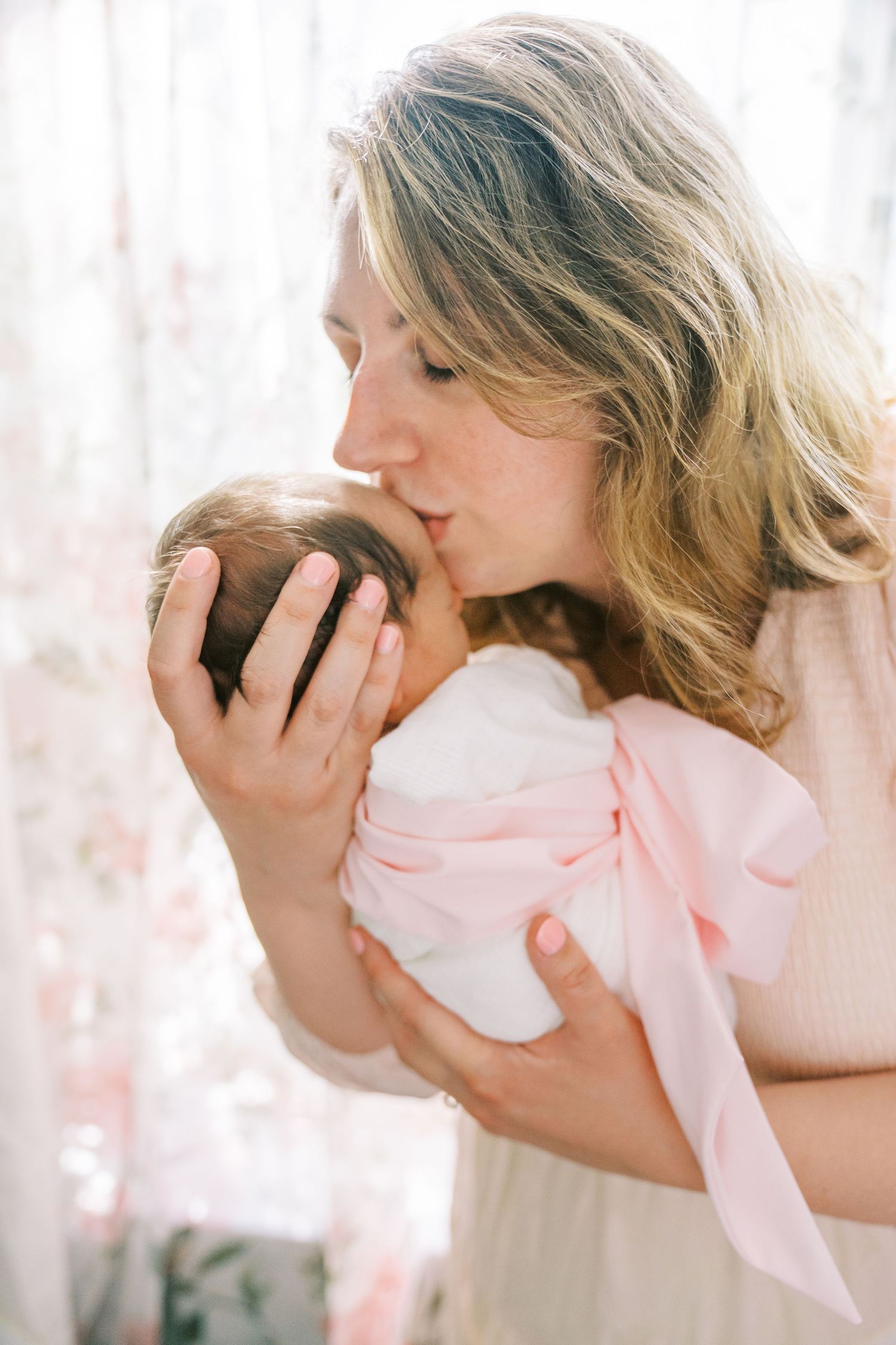 An intimate moment as mother kisses newborn wrapped in pink bow blanket by sheer curtains.