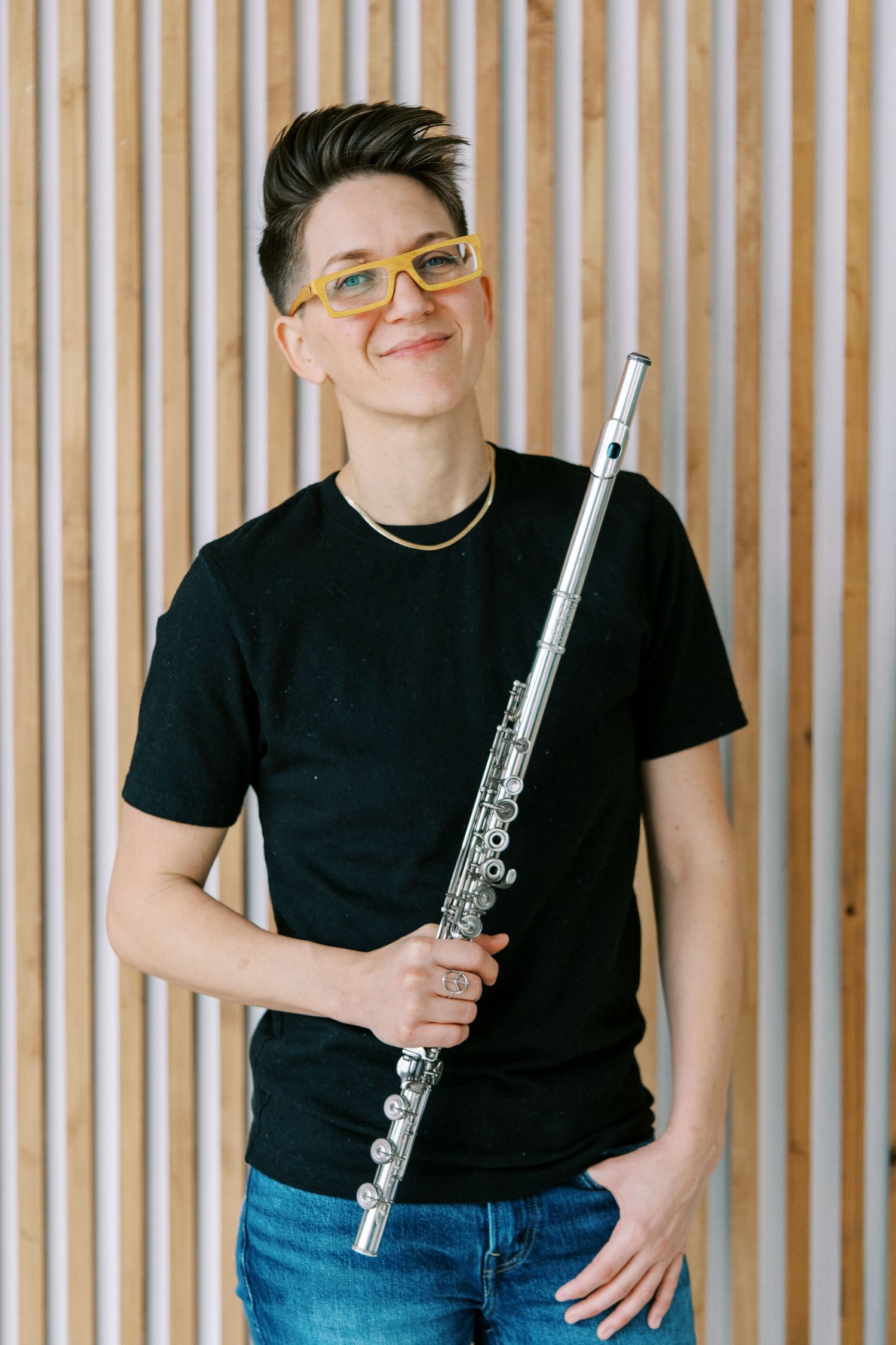A flutist in casual attire stands against a wooden slatted backdrop while holding their instrument.