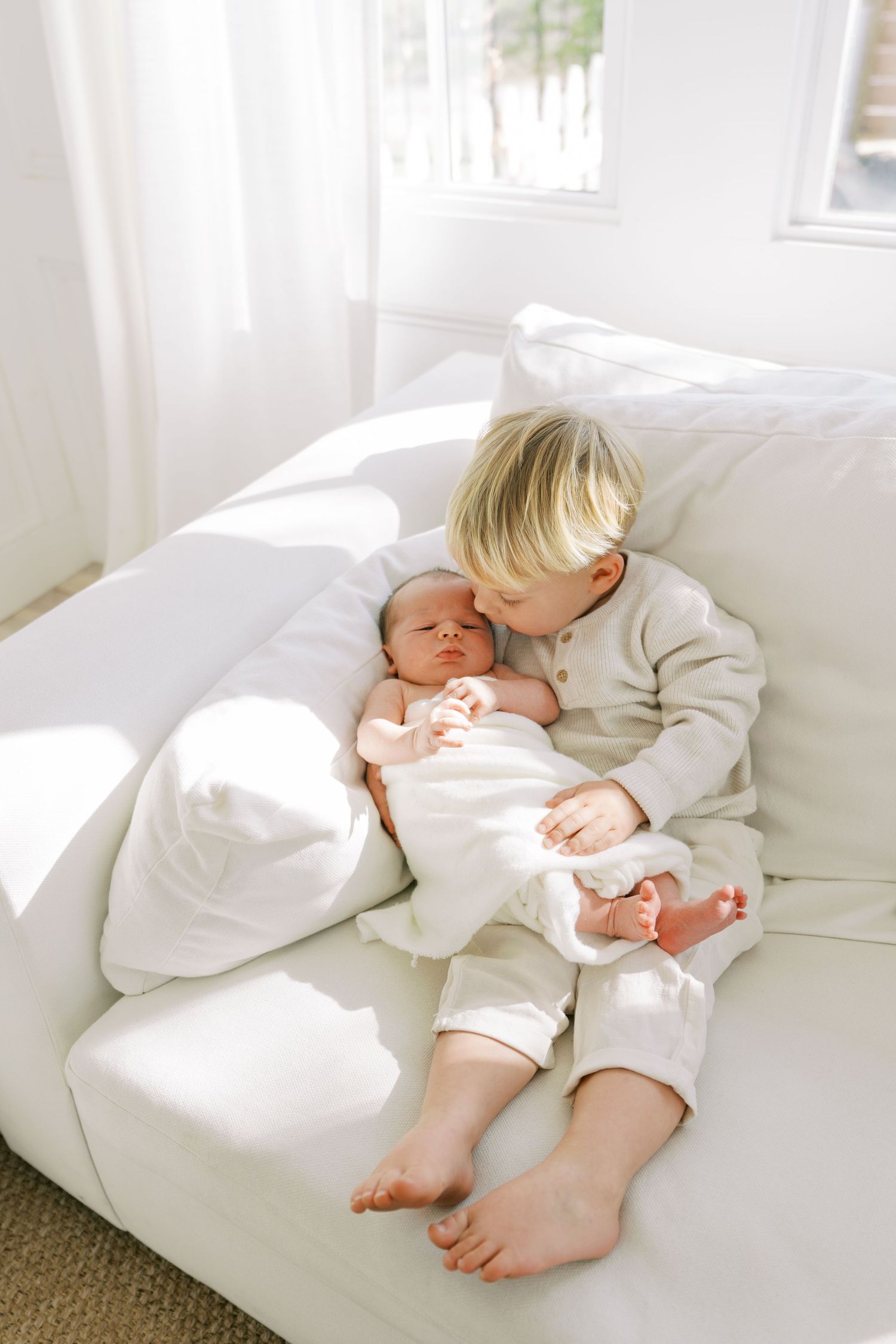 Two children in white clothing resting together on a bright white couch in a sunlit room.