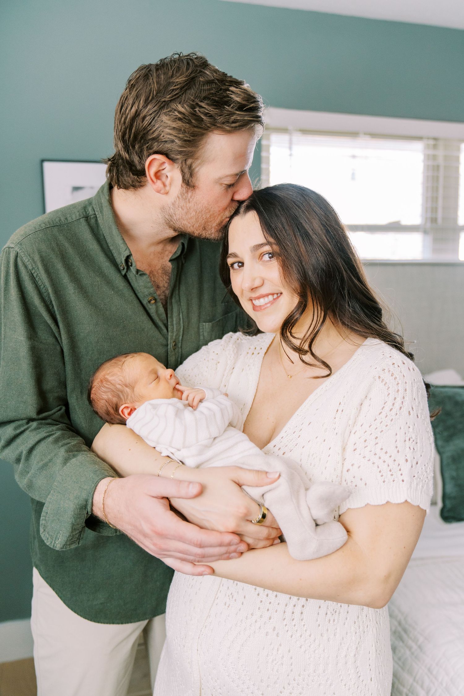A couple in green and white clothing shares tender moments with their newborn baby in a bright interior setting.