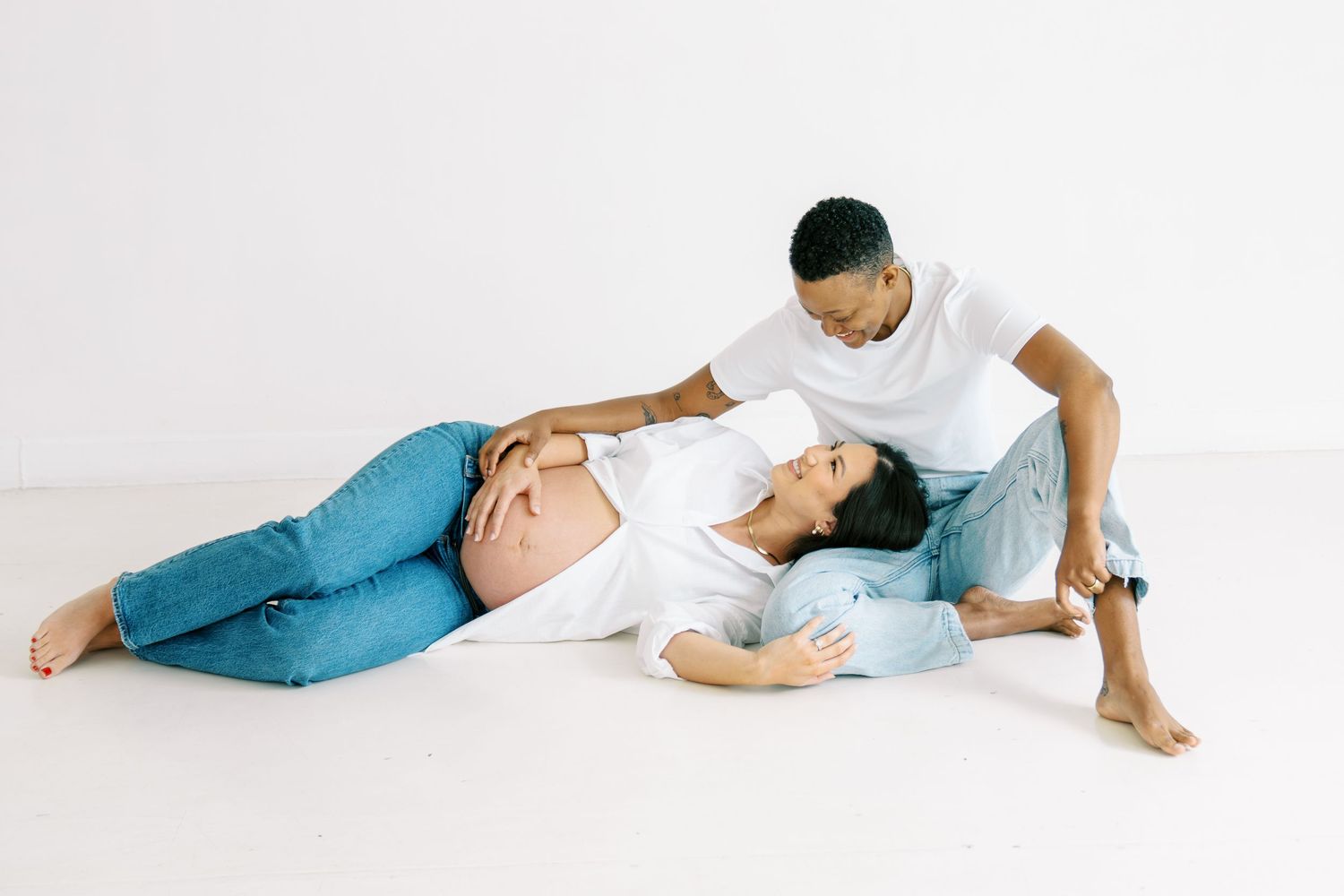 Pregnant woman lying on floor with partner sitting beside her, both wearing white tops and jeans in a bright studio.