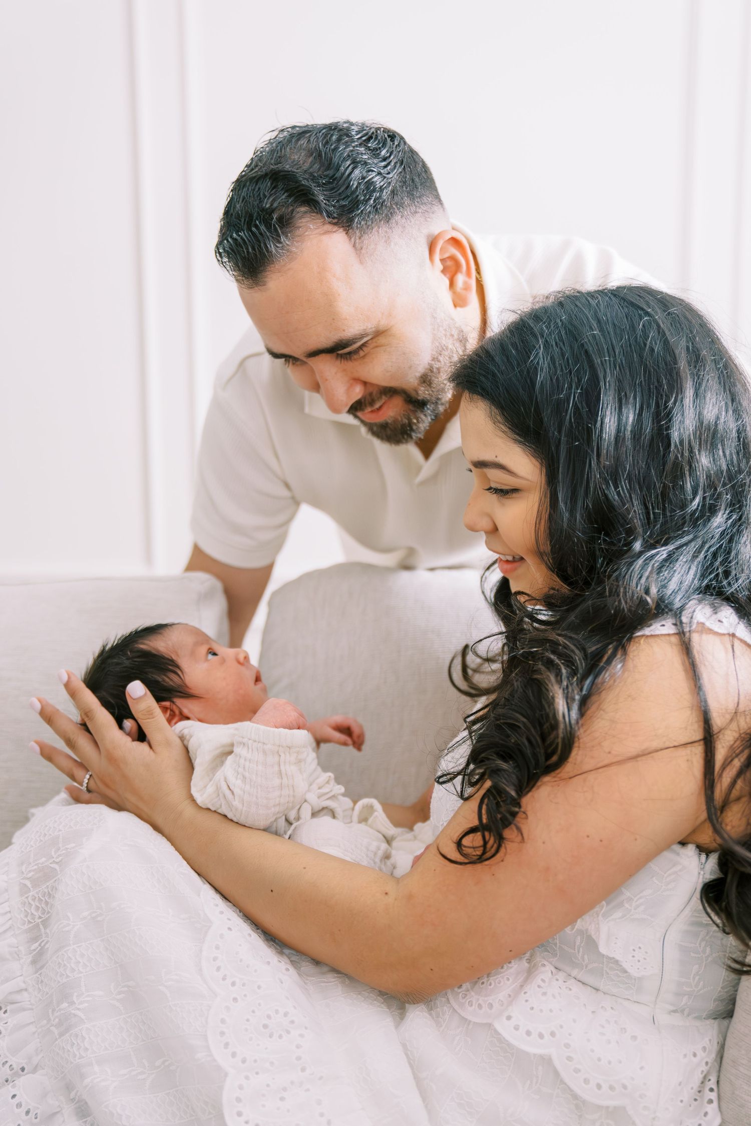 New parents lovingly gaze at their newborn baby, dressed in white, in a bright and airy nursery setting.