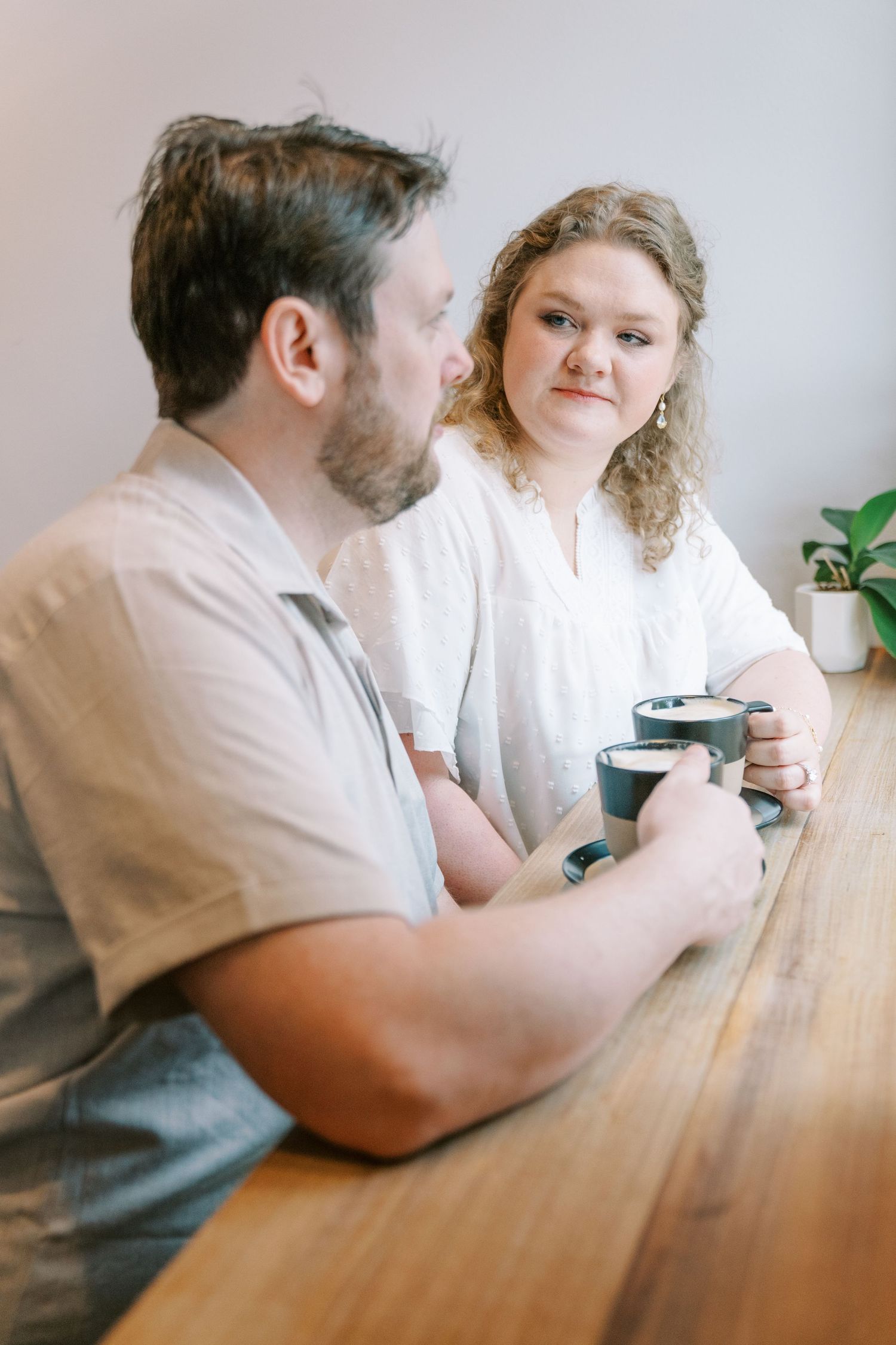 Bearded man and curly-haired woman sitting at a wooden bar counter sharing coffee cups in a bright cafe.