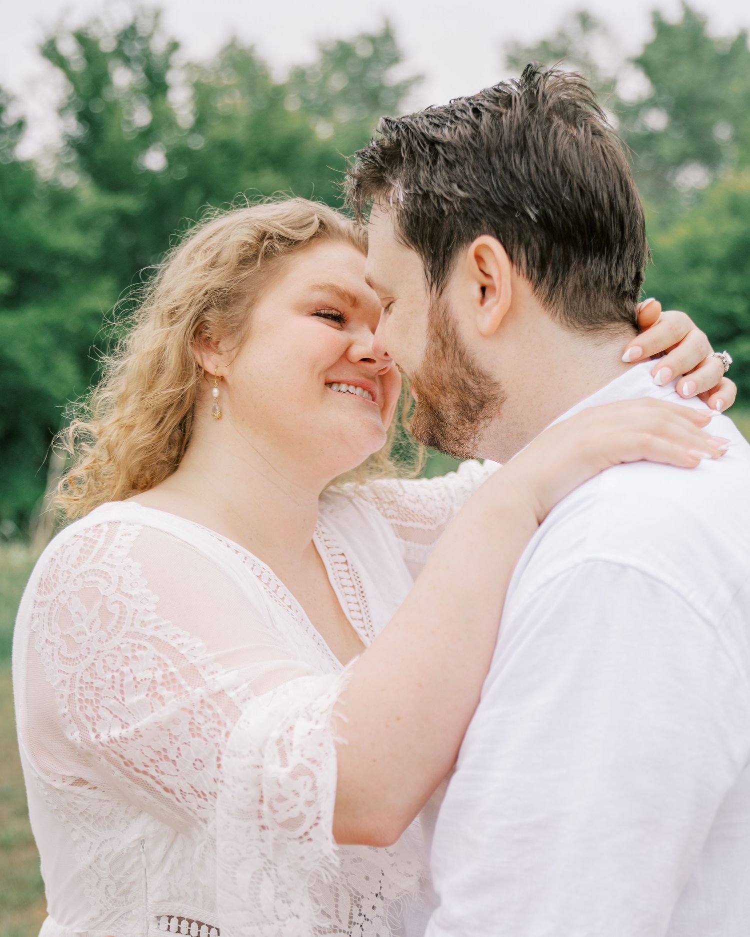 Smiling couple in white outfits share a romantic moment outdoors, touching foreheads with green trees in background.