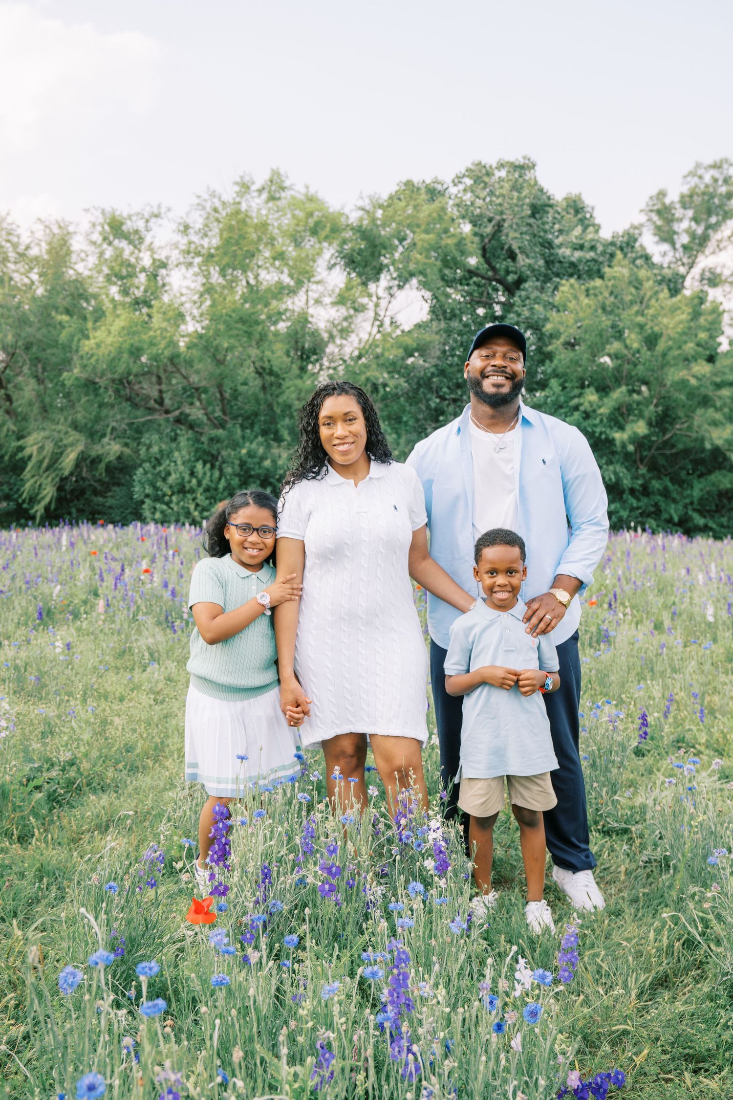 A joyful family of four poses in a wildflower field, wearing coordinated pastel outfits, surrounded by lush greenery.