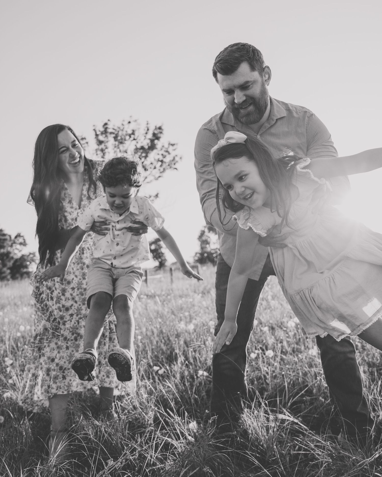 Black and white photo of a happy family playing in a sunny field, parents laughing with two young children.